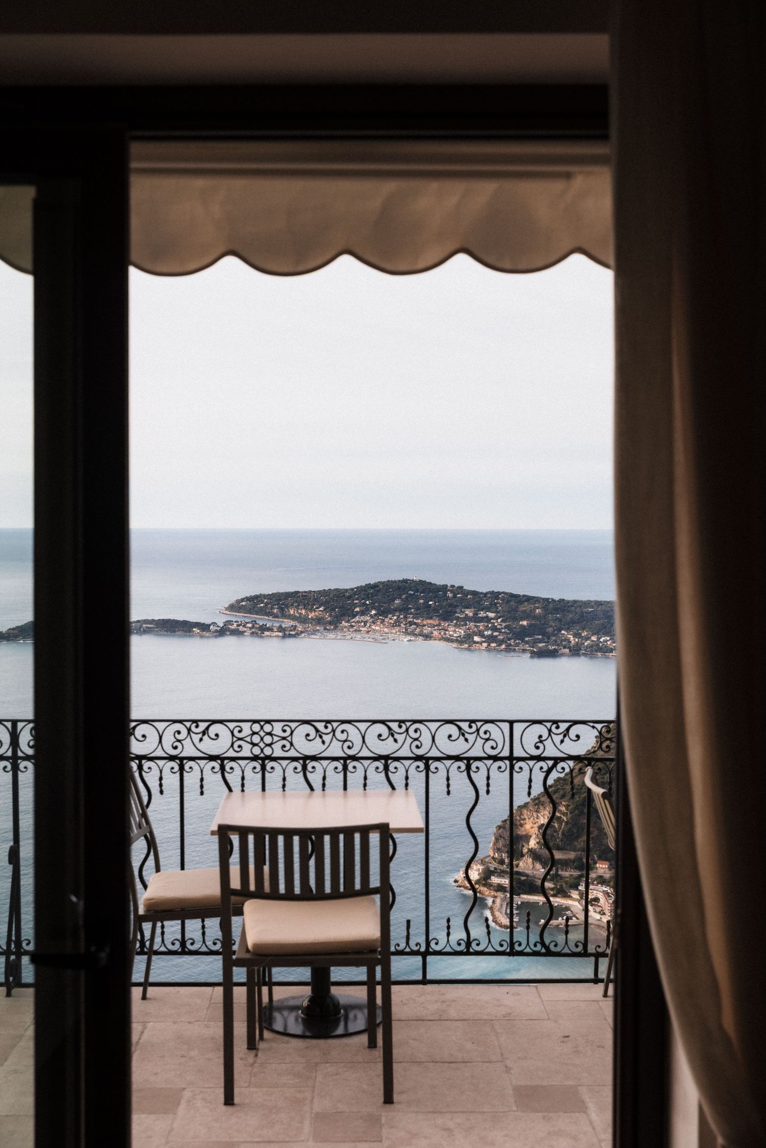 Château Eza - Elegant balcony with wrought iron railing, bistro table and chair, overlooking a serene Mediterranean sea and island view.