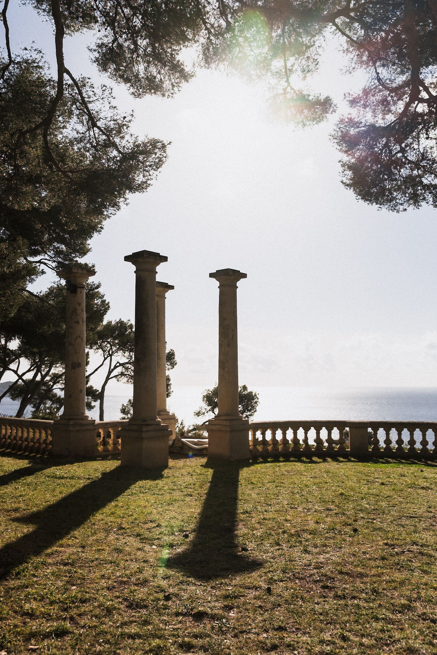 Villa Rocabella - Stone columns and balustrade terrace overlooking the sea, framed by pine trees in sunlight.