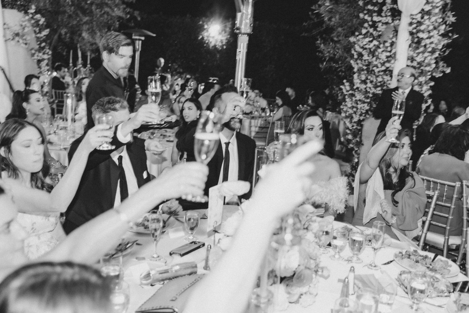 Guests raising champagne glasses in a toast at an elegant outdoor wedding reception dinner at night.