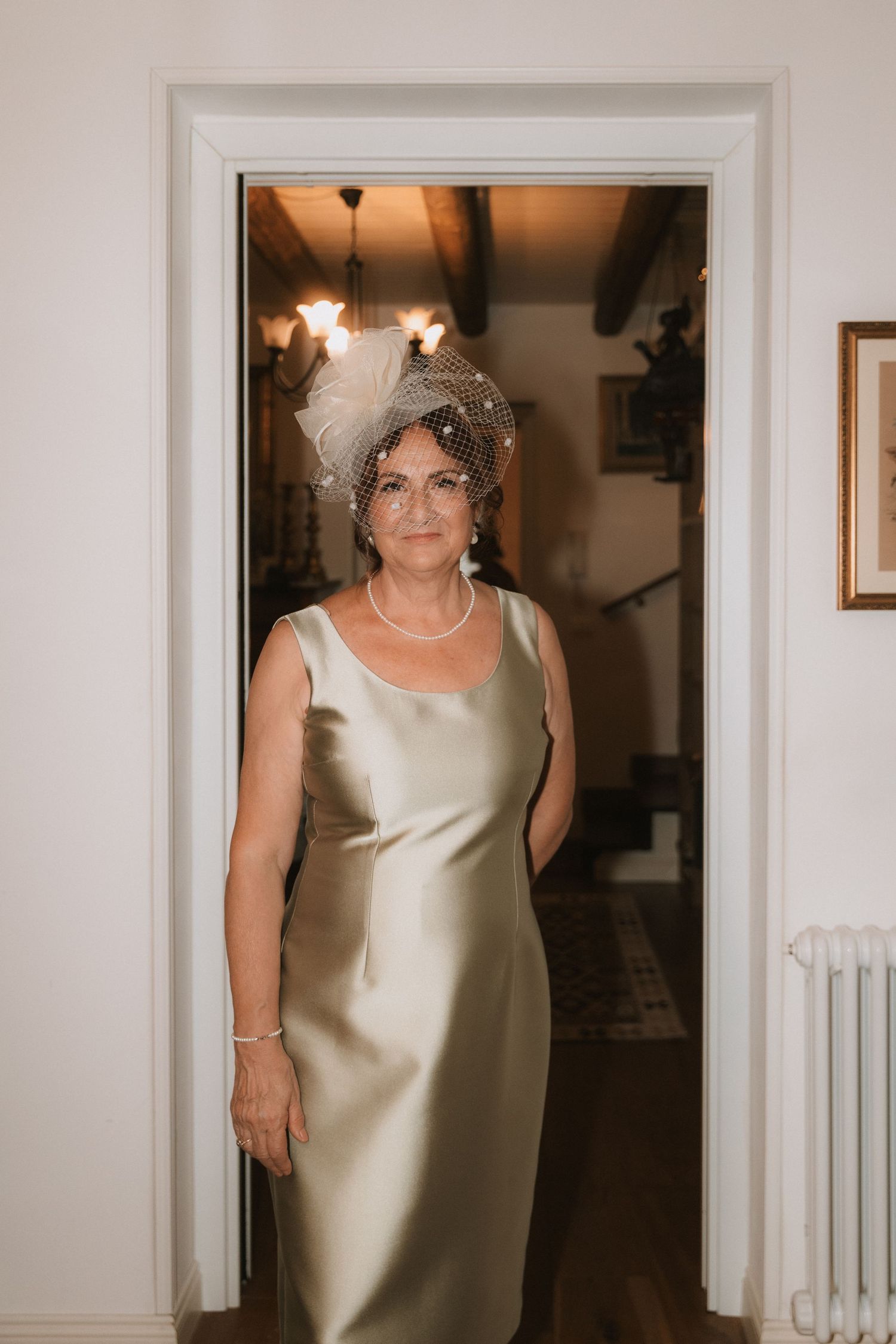 A woman in an elegant champagne-colored sleeveless dress poses in a white hallway interior.