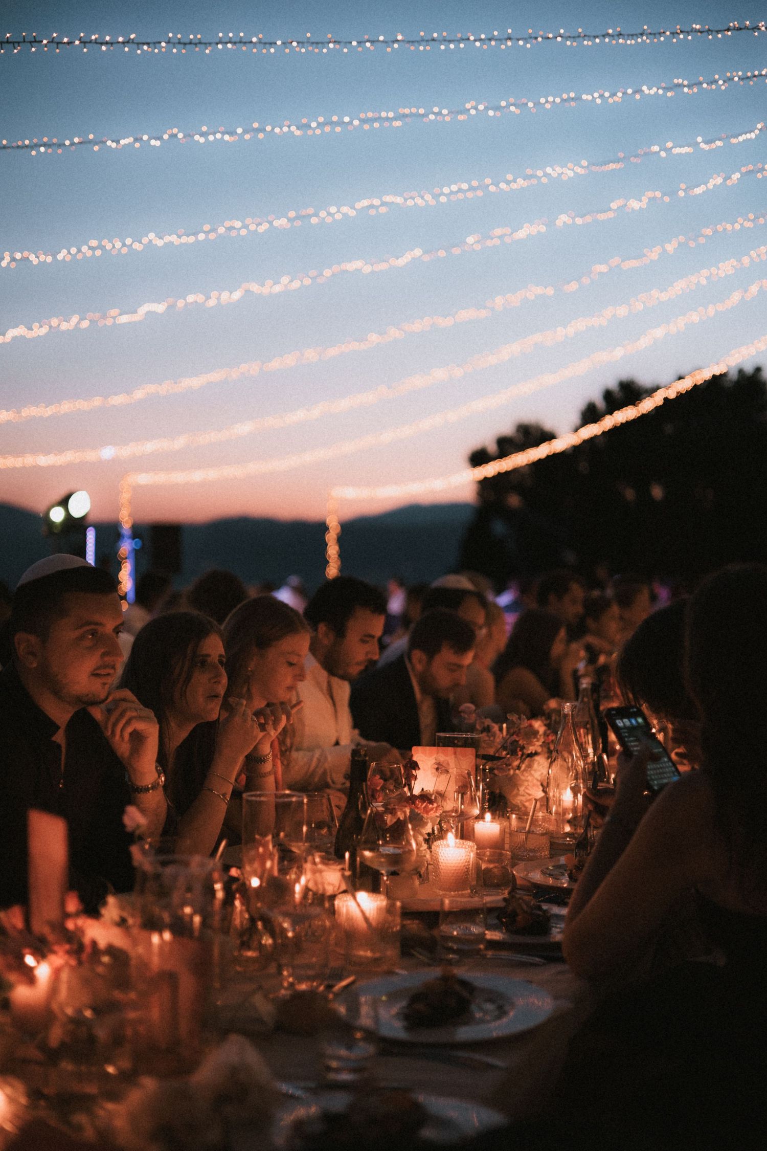 Outdoor dinner party at sunset with string lights overhead and glowing candles on long tables.