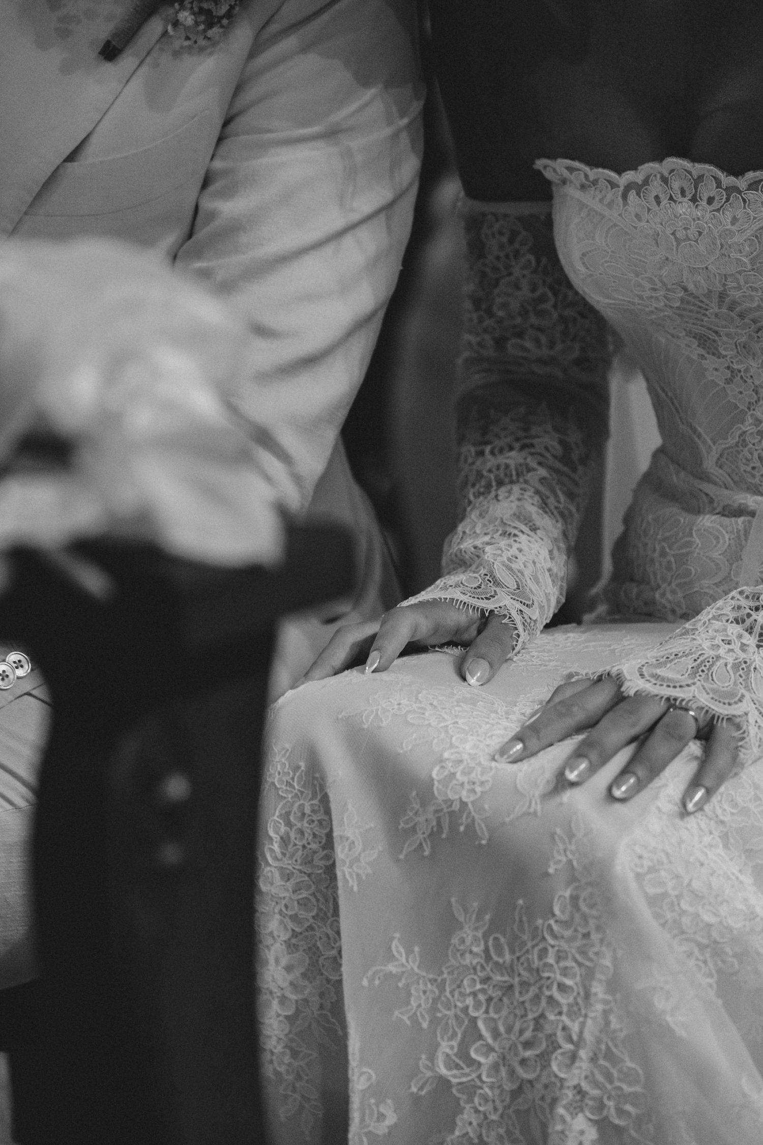 Black and white photo of bride and groom hands resting on lace wedding dress during ceremony.