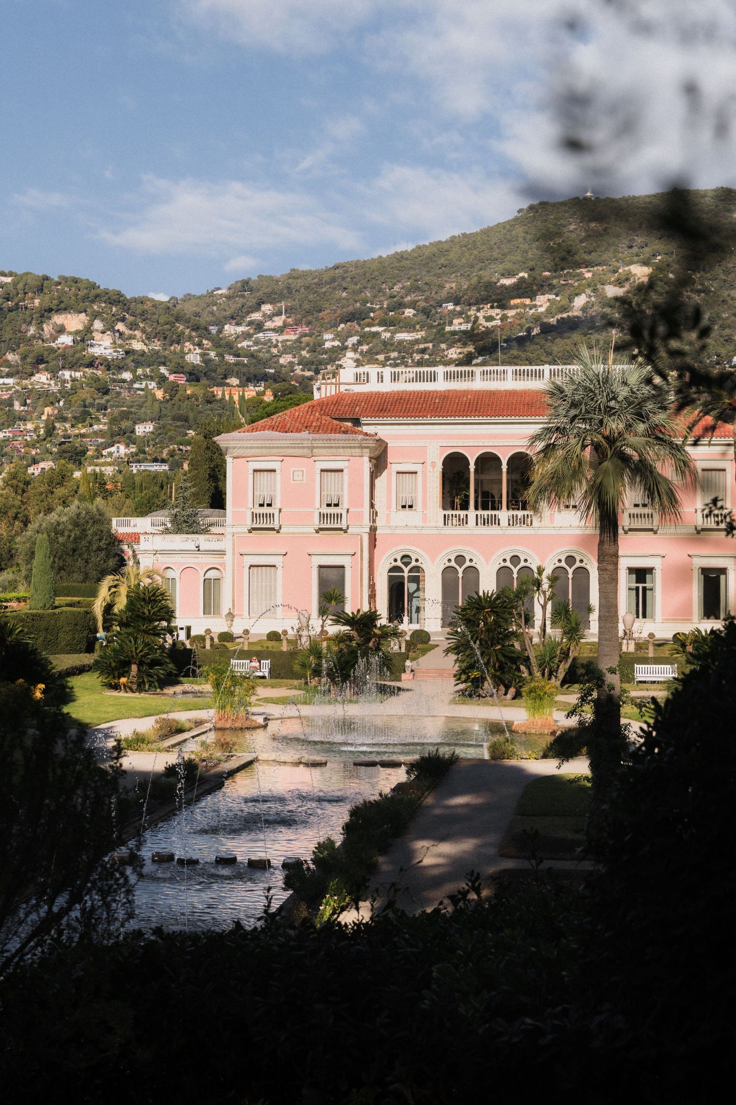 Villa Ephrussi de Rothschild - Pink villa with fountain garden, palm trees, and green hills in the French Riviera under a blue sky.