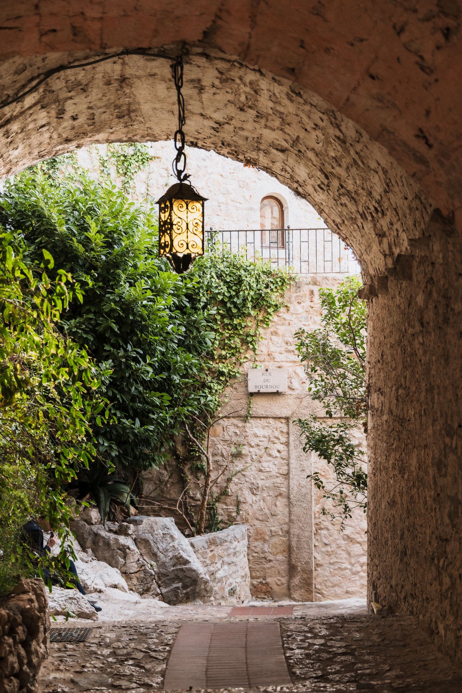 Château Eza - Stone archway with ornate hanging lantern leading to a lush courtyard in a medieval village.