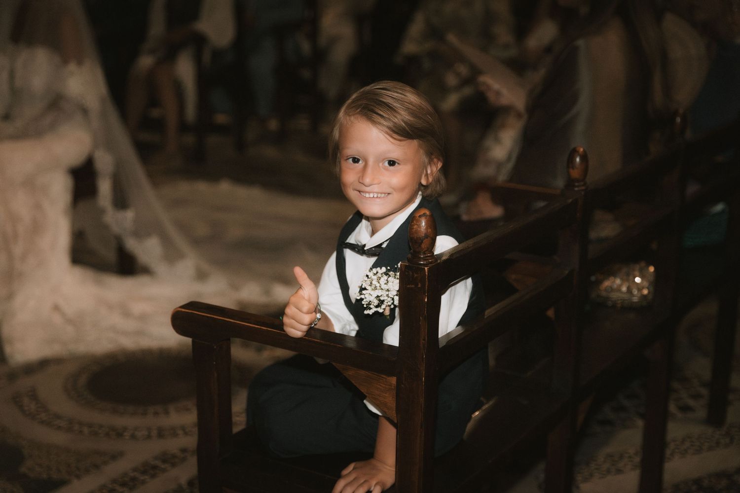Young ring bearer in suit gives thumbs up at wedding ceremony, sitting in wooden pew with boutonniere.
