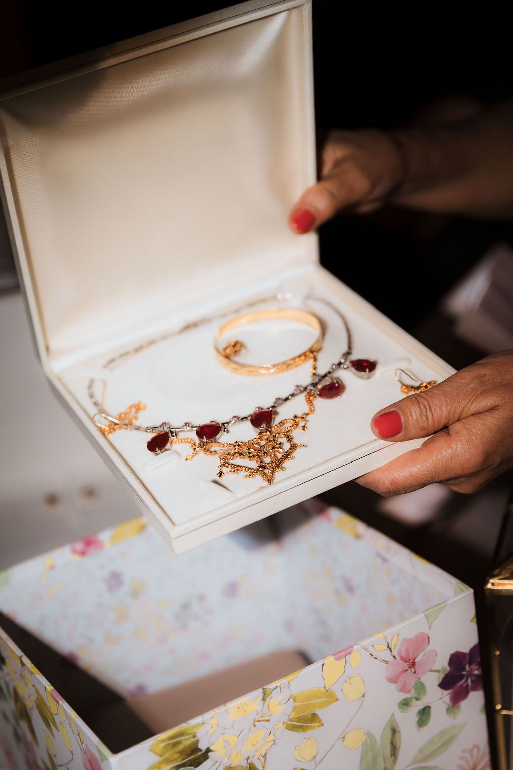 Hands opening a decorative box revealing gold jewelry with red accents against a floral patterned background.
