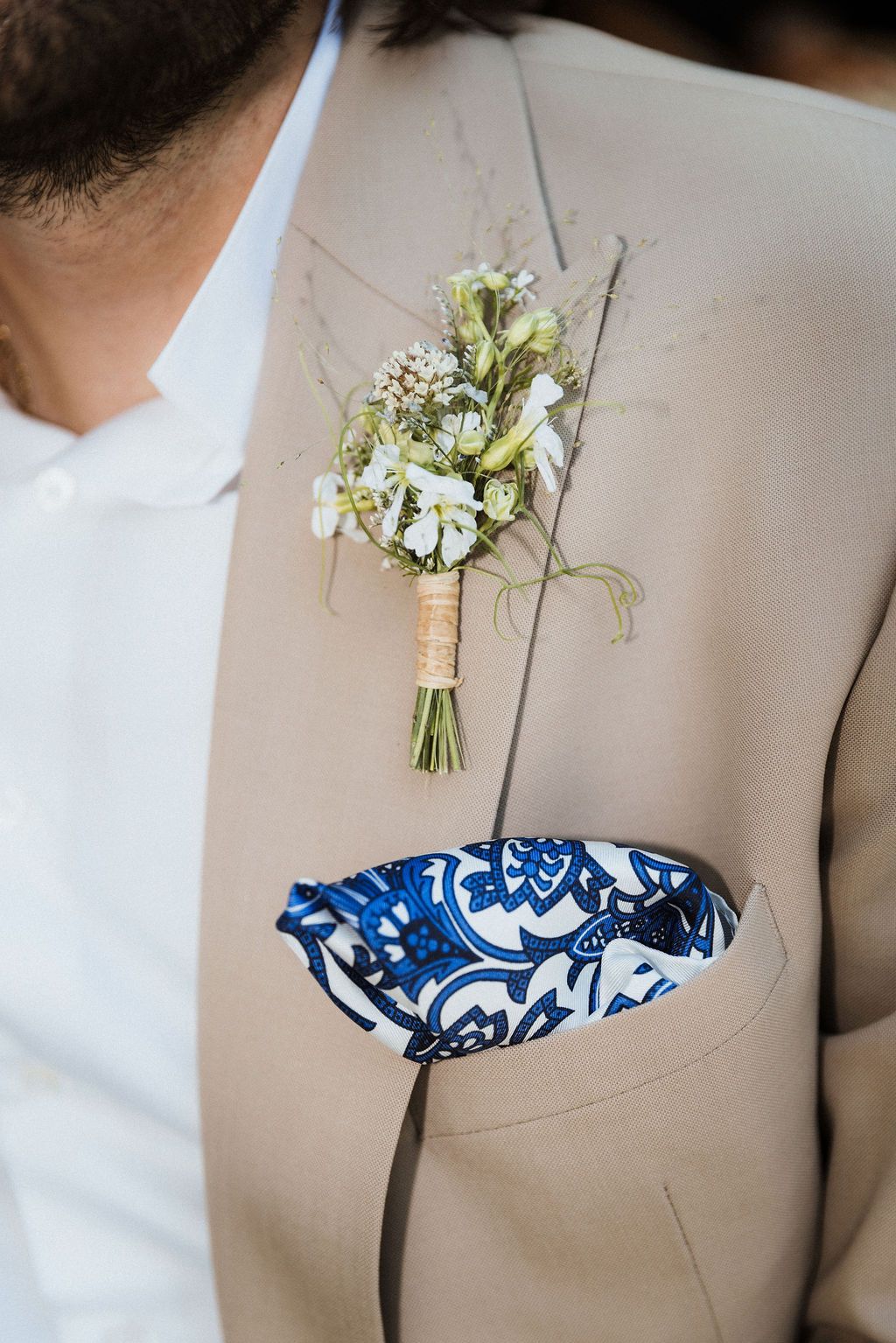 Close-up of a beige suit jacket with a blue patterned pocket square and delicate white floral boutonniere.