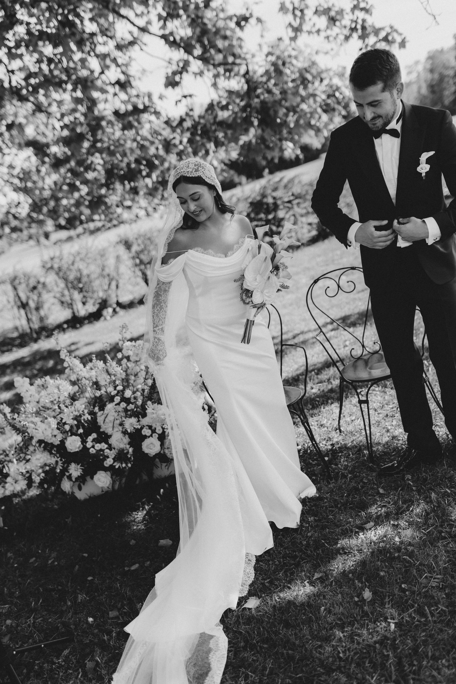 Elegant couple in wedding attire stand together in a garden setting with decorative iron bench.