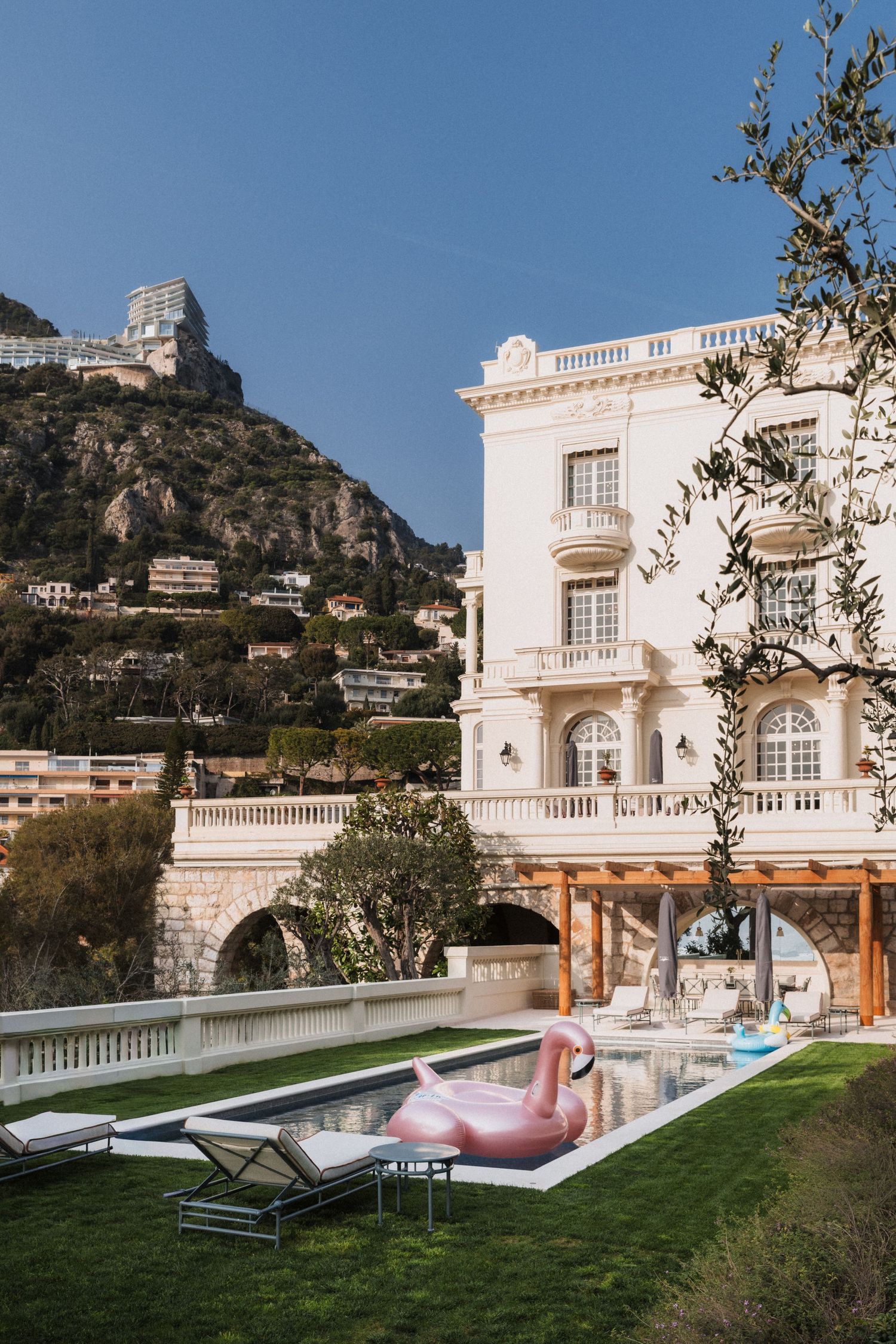 Villa La Vigie, Monaco - Elegant white villa with balconies, lush garden, pool with pink flamingo float, and scenic rocky hillside backdrop.