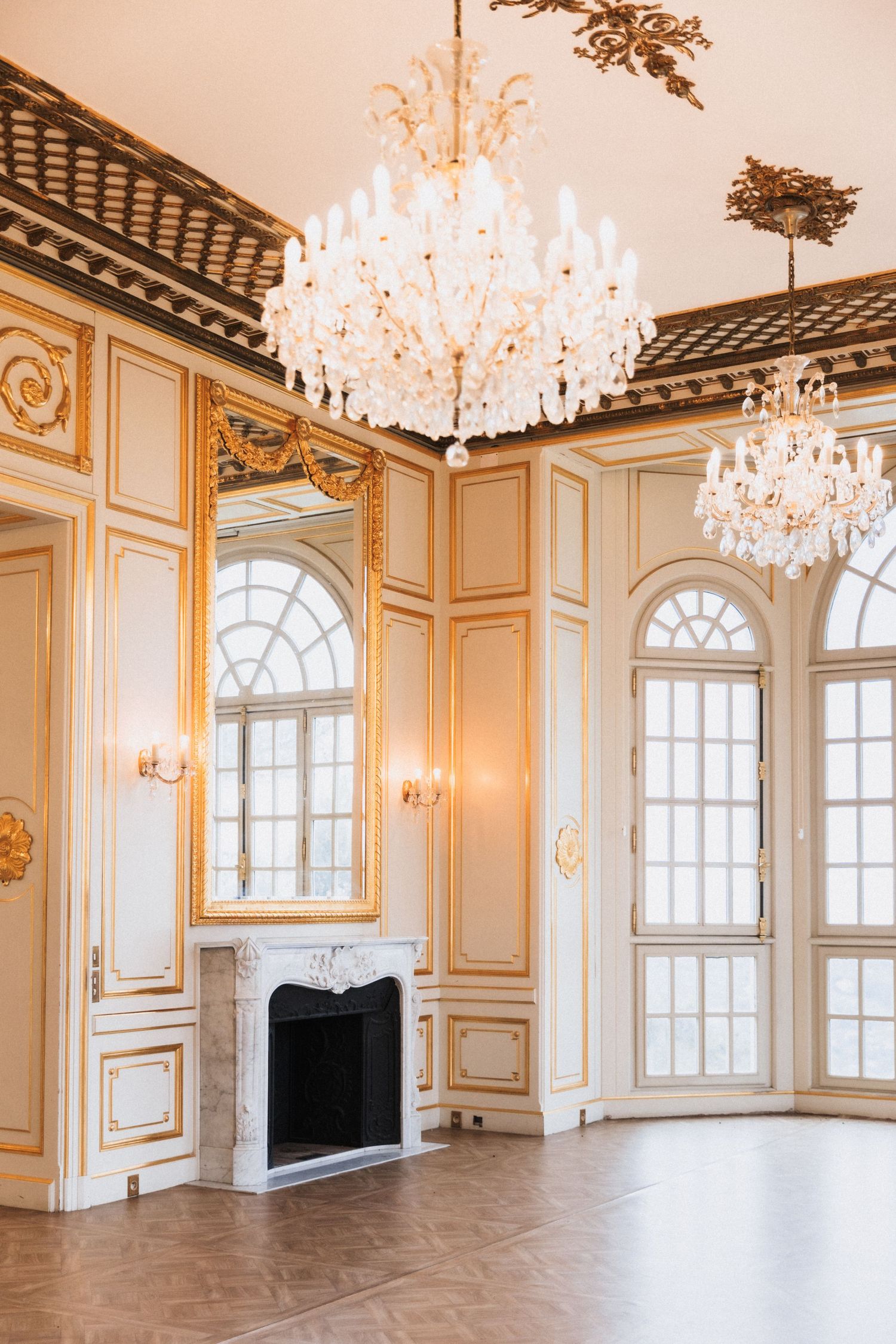 Elegant ballroom interior with crystal chandeliers, ornate gold trim, arched windows, and white walls in classic French style.