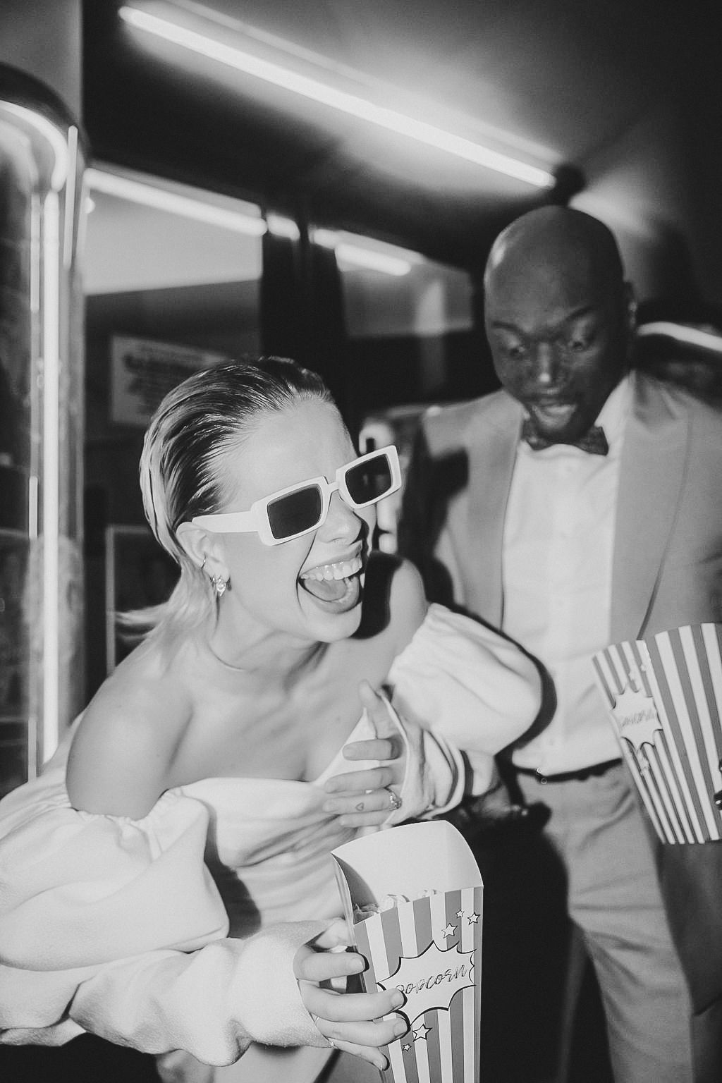 Black and white photo of a laughing bride in sunglasses holding popcorn with a man in a suit at a cinema-themed wedding.