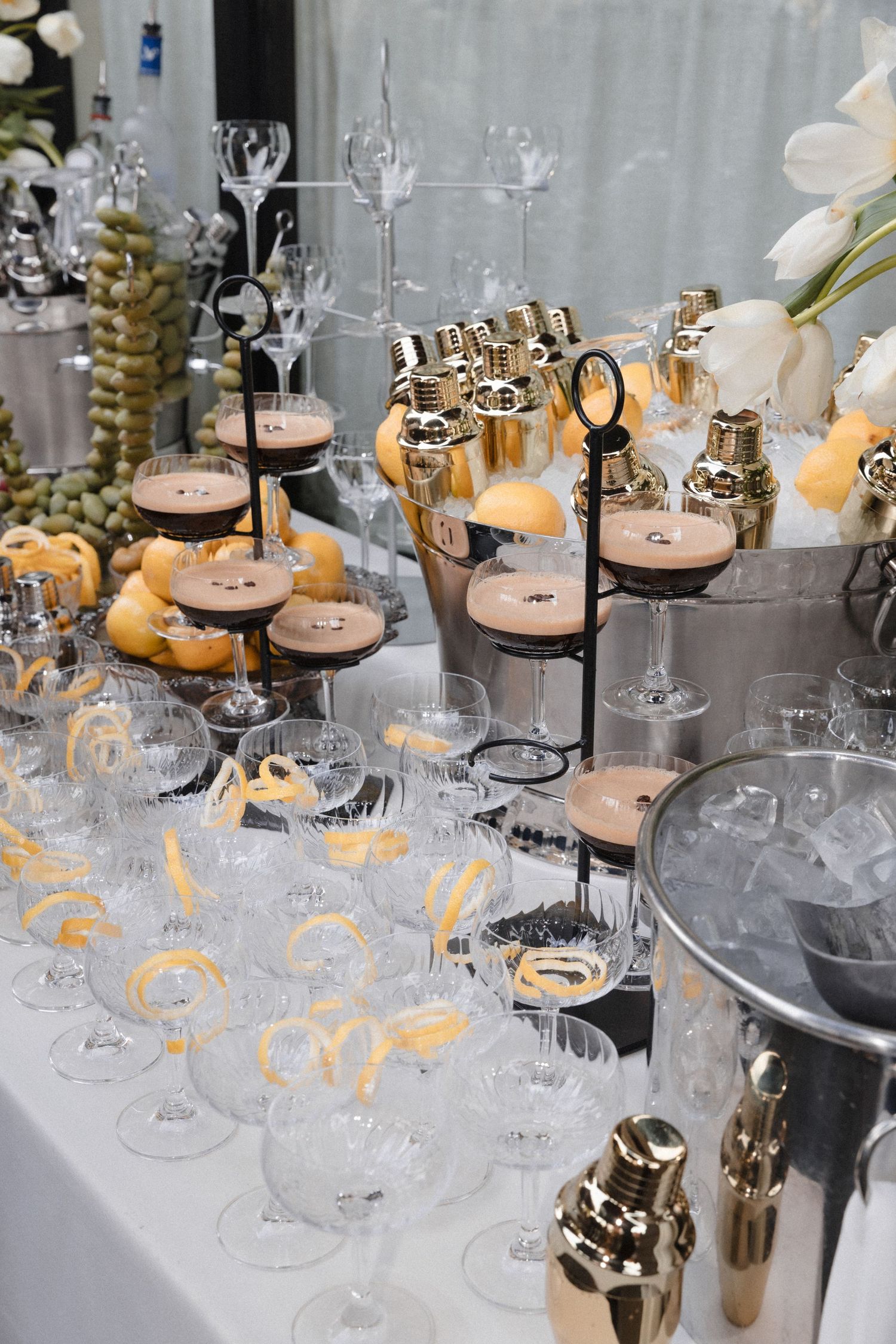 Elegant wedding cocktail bar display with champagne glasses, gold shakers, and floral arrangements on a white table.