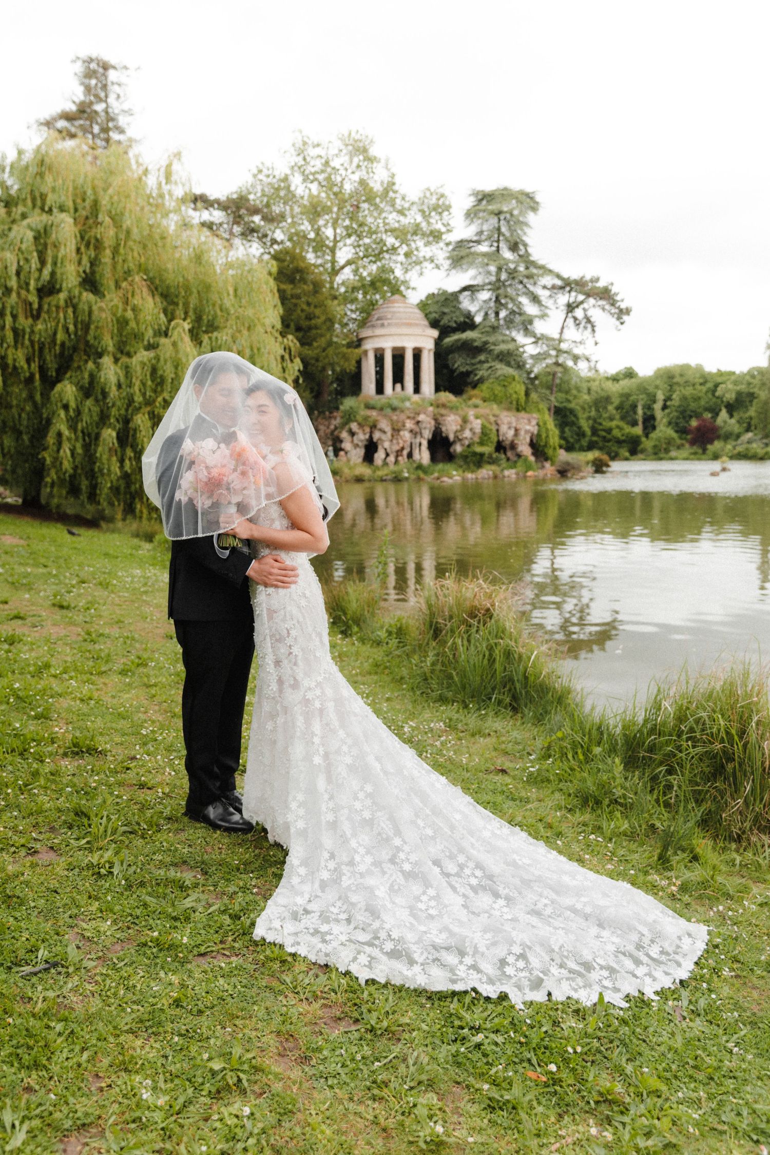 Bride and groom share a romantic moment under a veil by a serene lake, with a classical gazebo in the background.