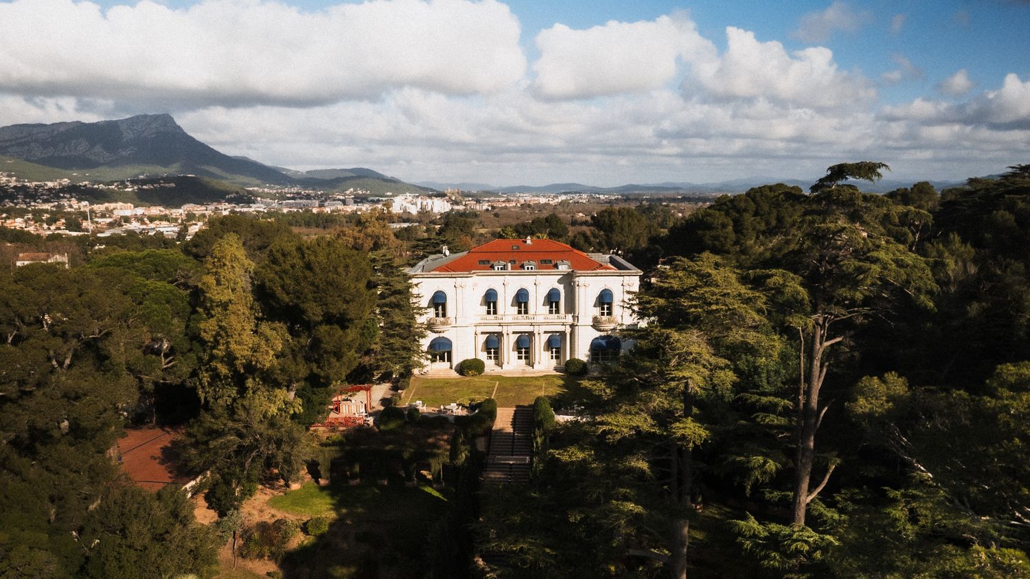 Aerial view of a grand white villa Rocabella with red roof surrounded by lush trees, with mountains and cityscape in background.