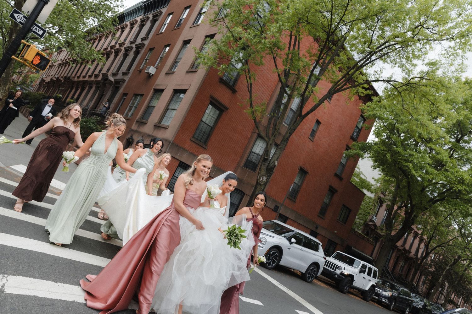 Bride and bridesmaids in elegant gowns crossing a city street with brick buildings and trees in the background.