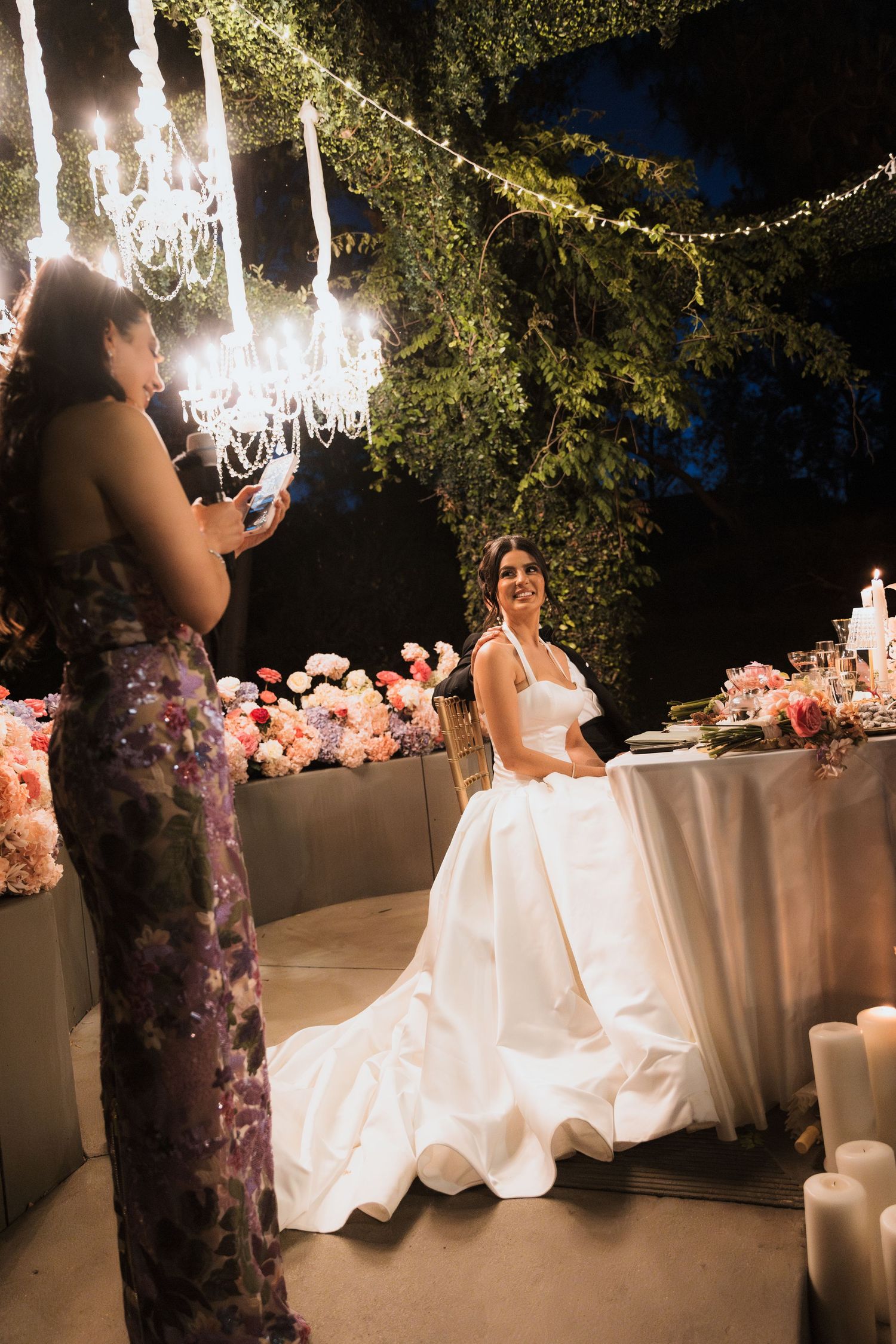 Bride in strapless white gown smiles at guest speech during elegant outdoor wedding reception.