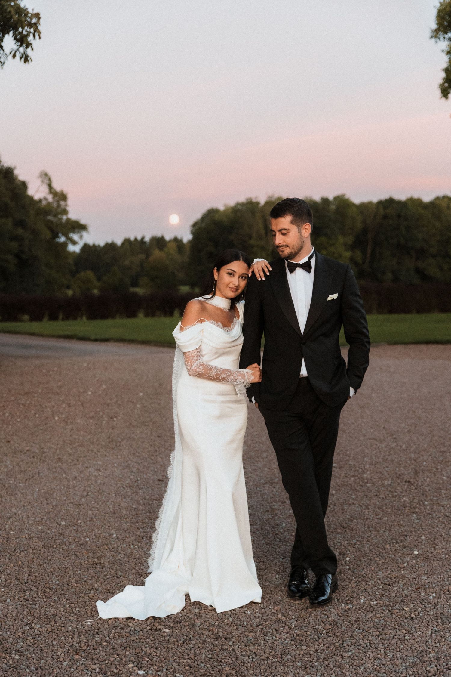 A romantic wedding couple walks together at sunset in an open field with a soft pink sky in the background.