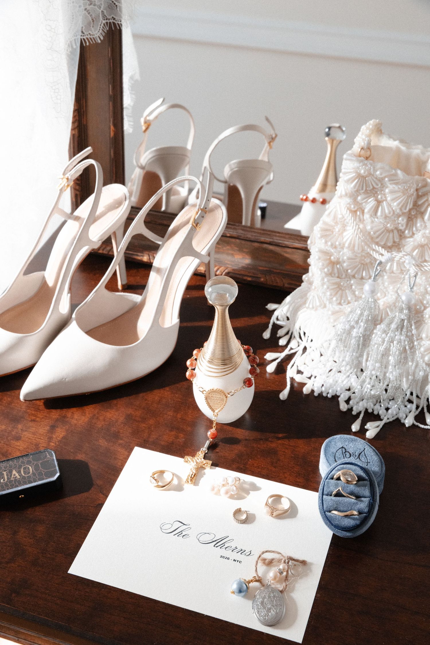 Elegant bridal accessories including white heels, Dior perfume, rings, and wedding stationery on table.