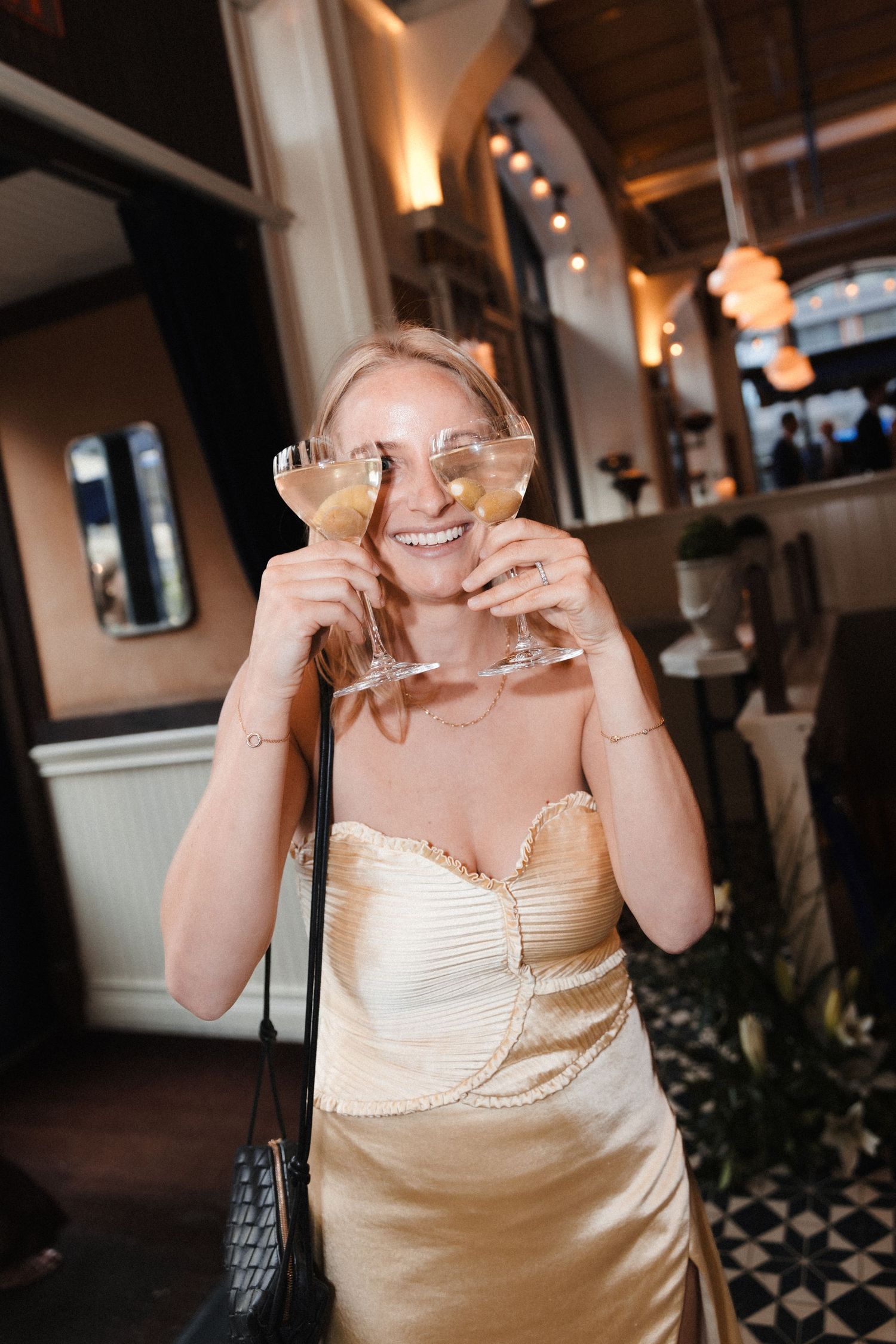 Smiling woman in gold strapless dress holding two champagne glasses in a stylish restaurant setting.