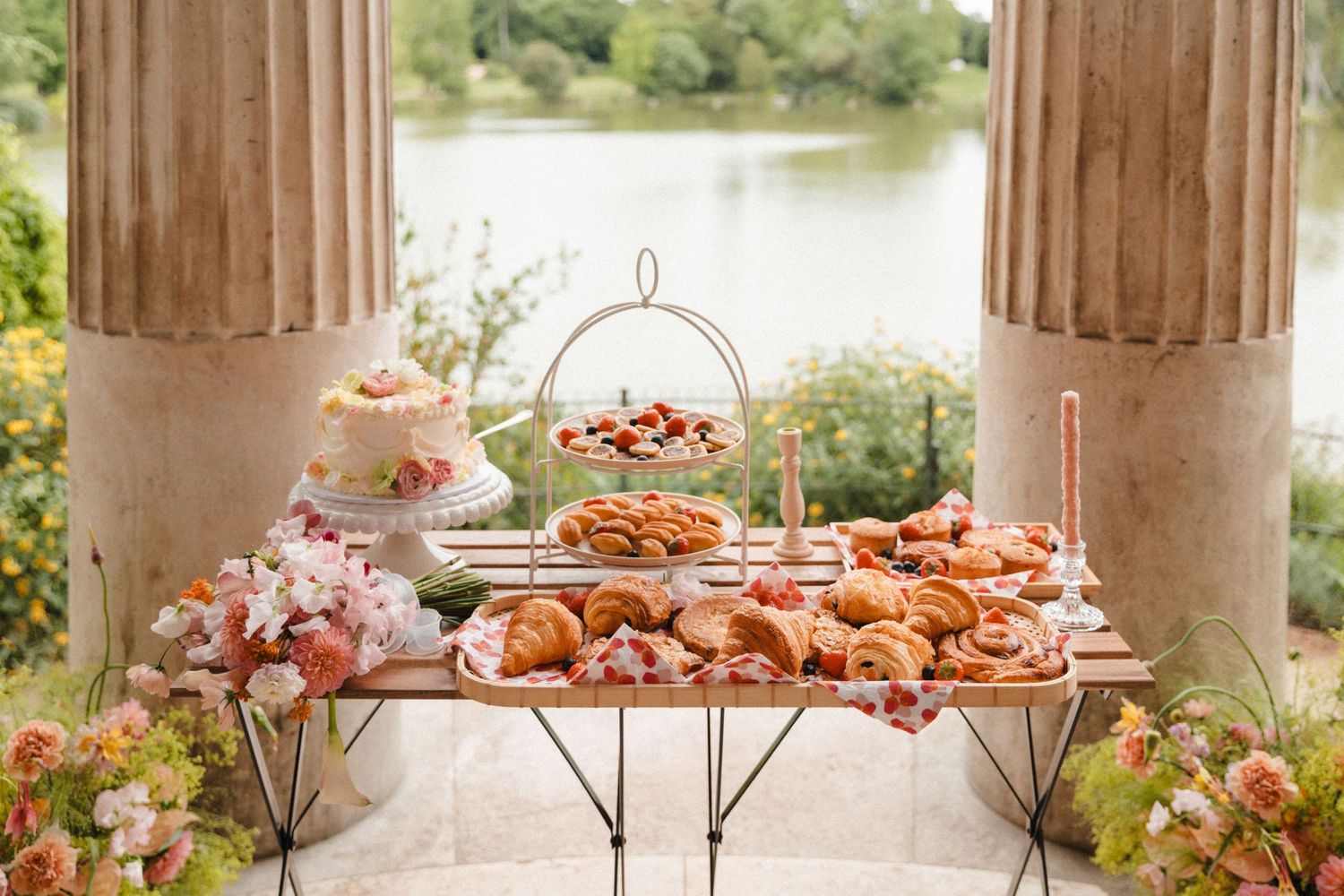 Elegant outdoor dessert table with pastries, cake, and strawberries between stone columns overlooking a serene lake.