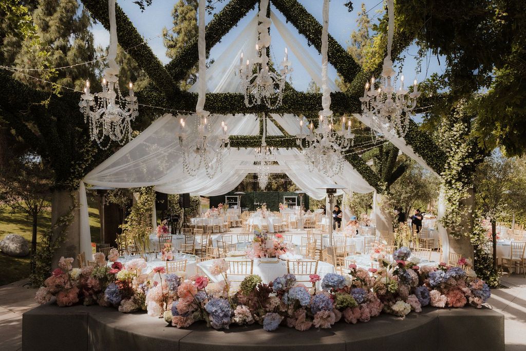 Outdoor wedding reception setup with floral arrangements and chandeliers hanging from trees.