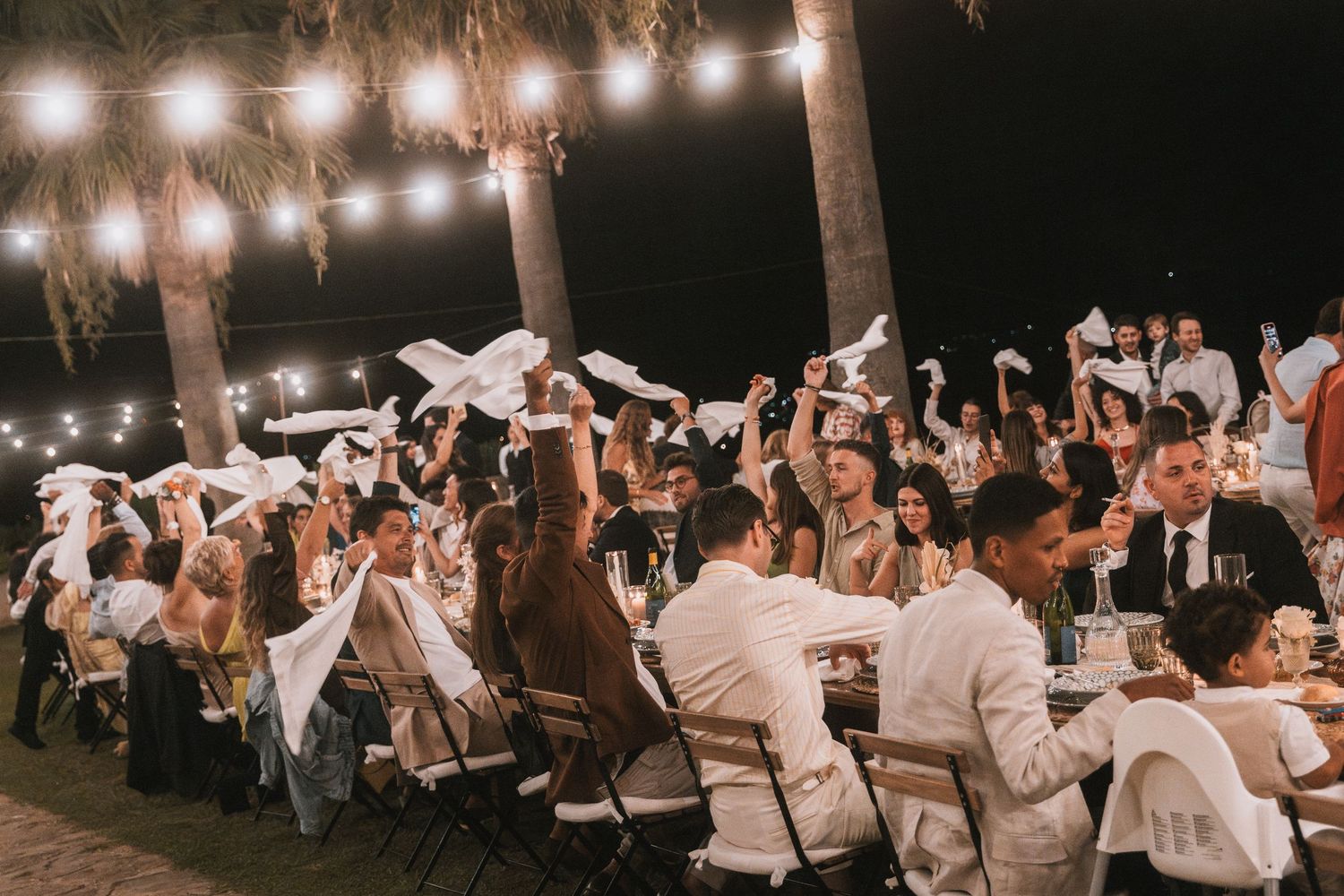 Outdoor evening wedding reception with string lights and celebrating guests.