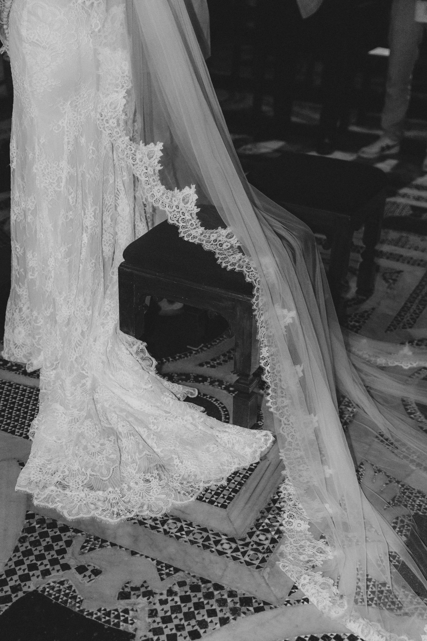 Black and white photo of a lace wedding dress and veil with intricate details laid out on a patterned floor.