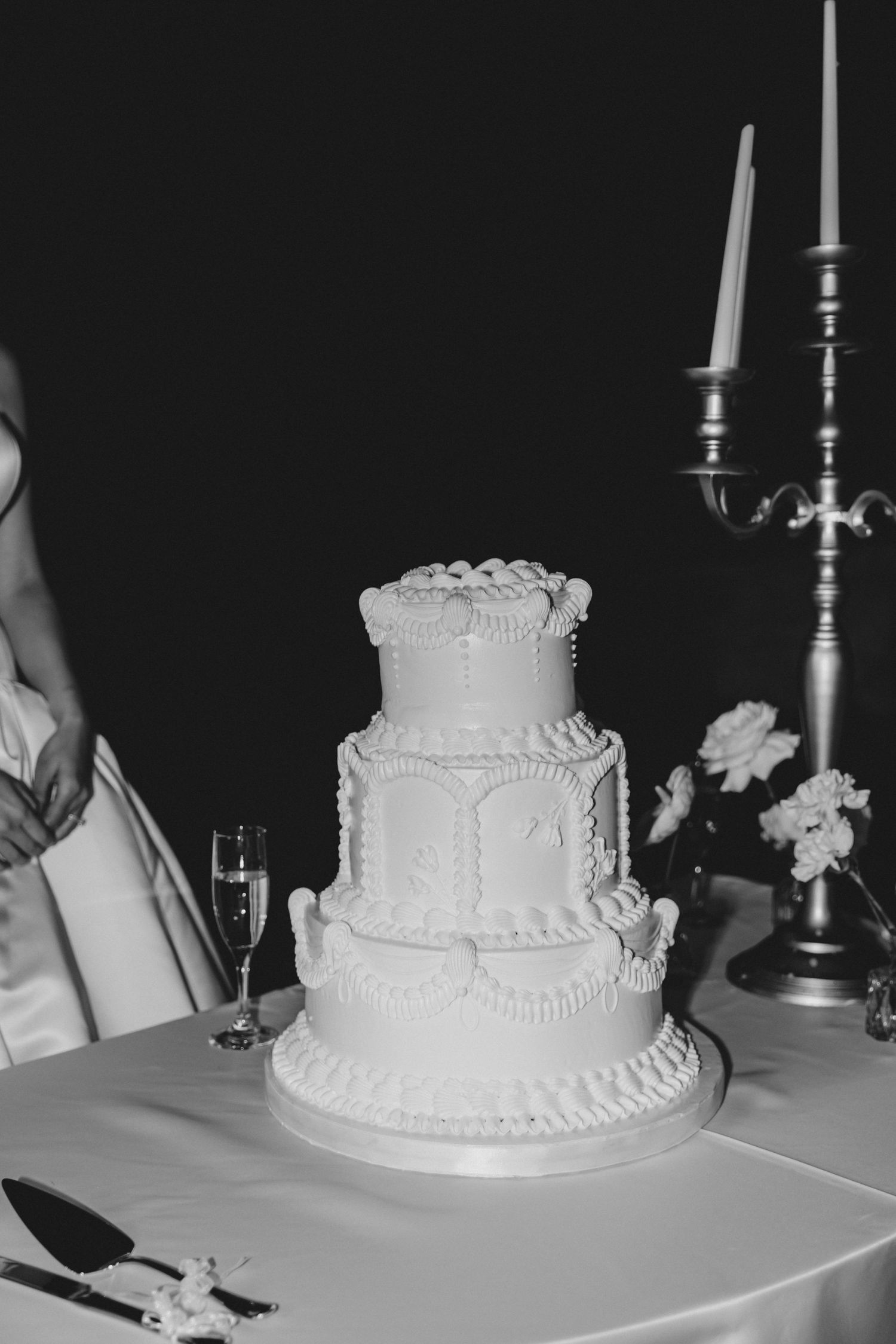 A classic white tiered wedding cake with delicate piping details sits elegantly on a table with candlesticks nearby.