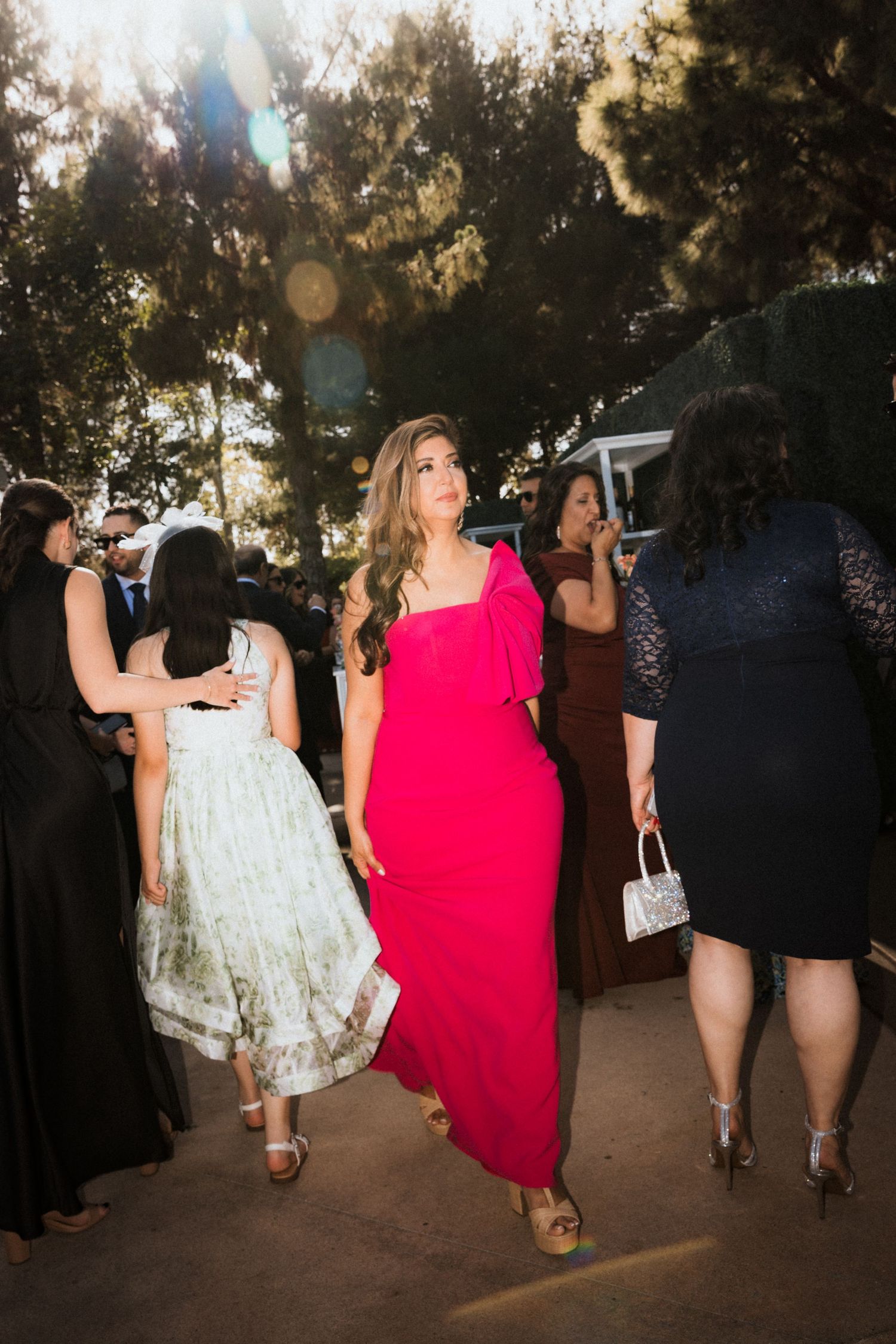 A group of elegantly dressed women walking together at an outdoor event with trees in the background at sunset.