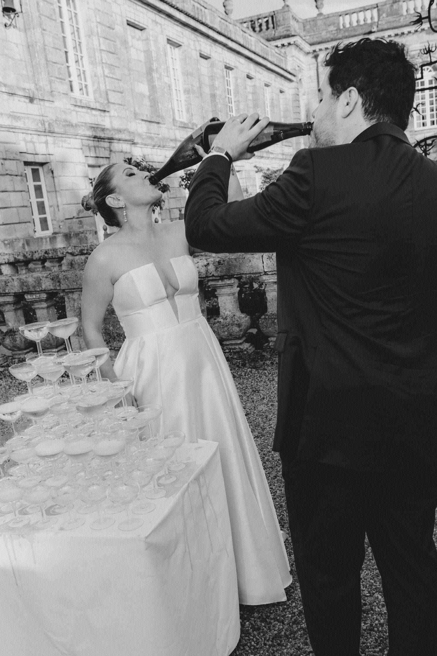 Newlyweds pour champagne into a tower of glasses at an elegant outdoor wedding reception.