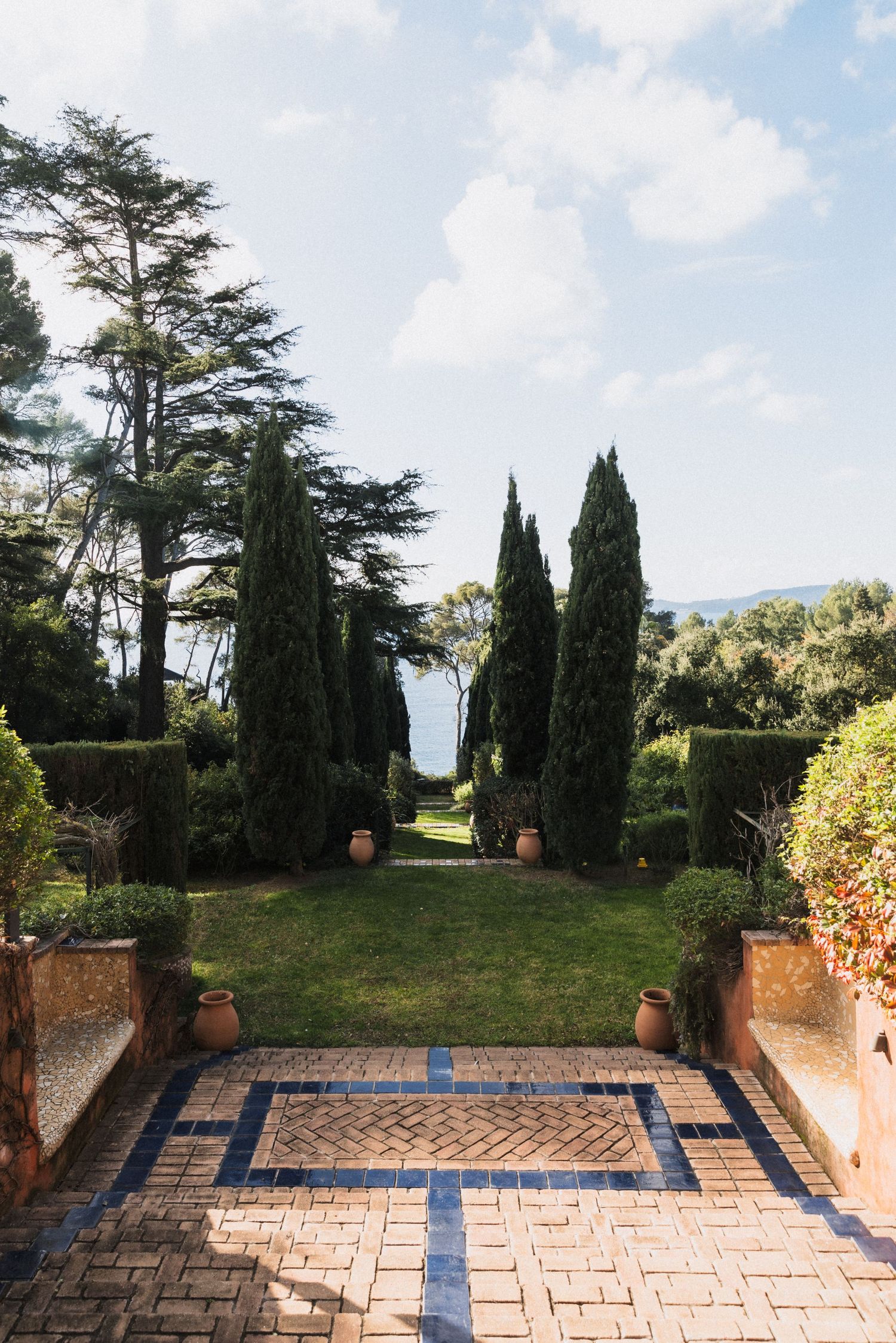 Villa Rocabella - Elegant Italian garden with cypress trees, & herringbone brick pathway leading to a scenic coastal view.
