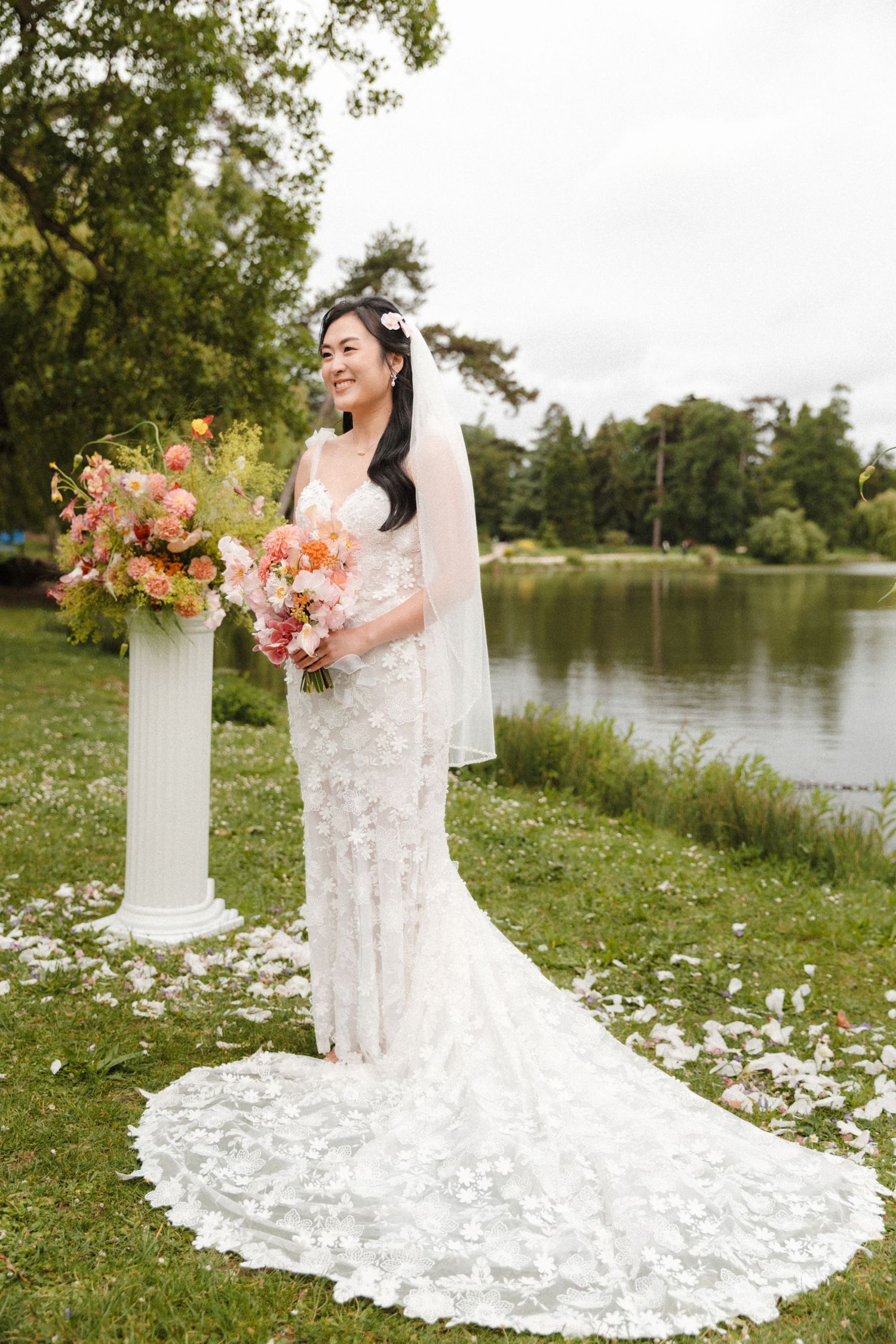 Bride in floral lace gown with veil holds pink bouquet by lakeside garden with white column floral arrangement.