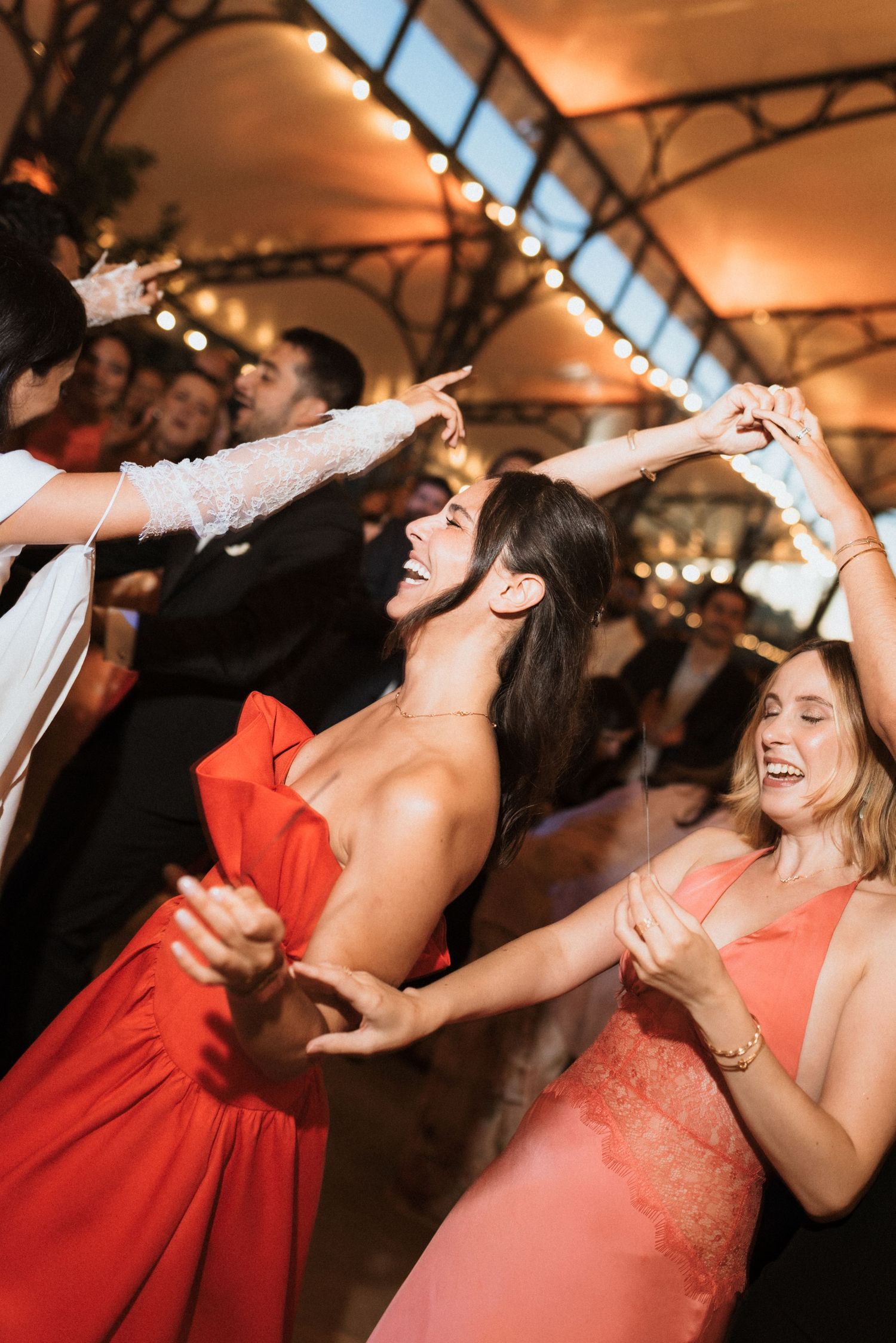 Wedding guests in red and coral dresses dance joyfully under warm lighting at an indoor reception.