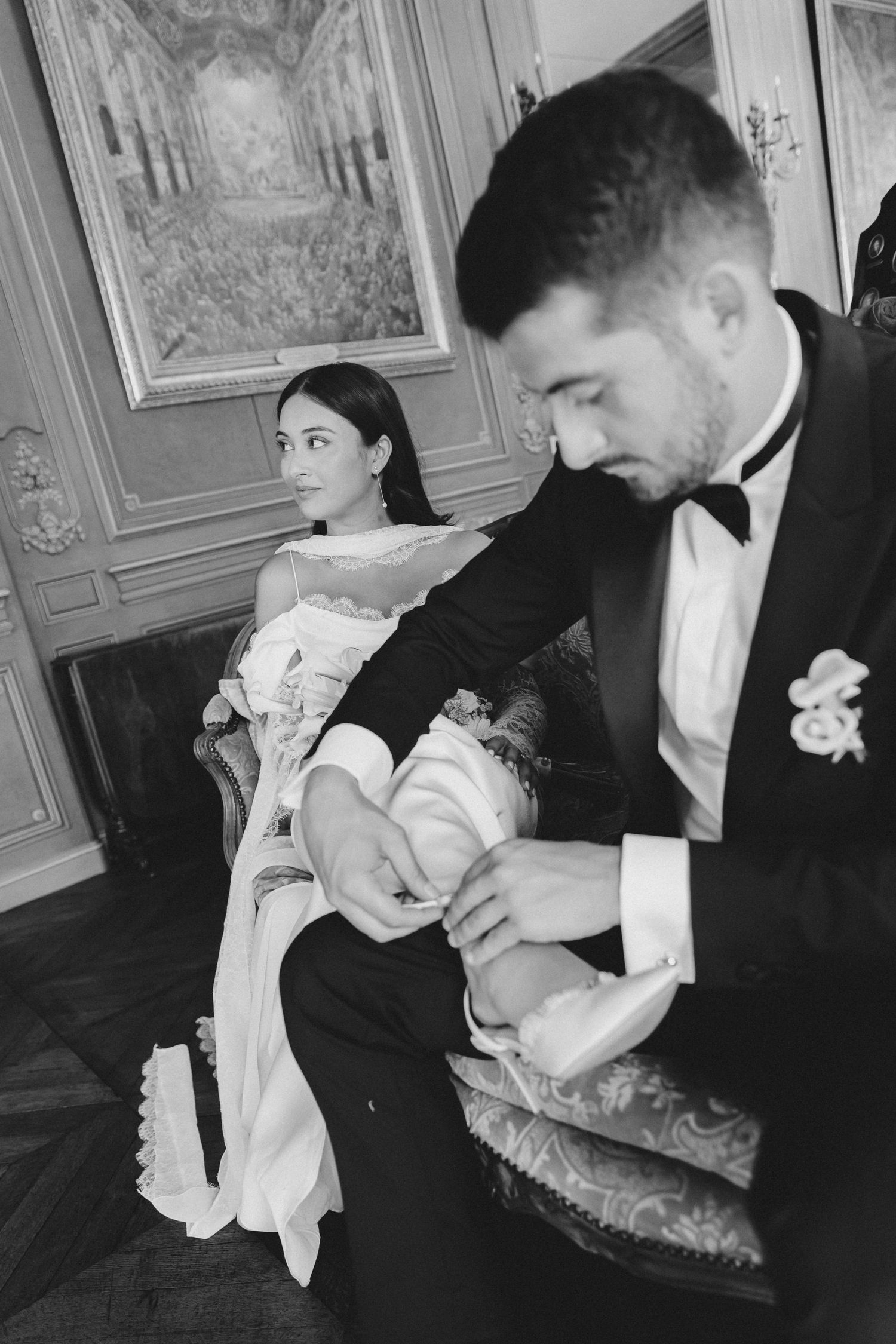 A couple in formal attire sits in an elegant room with ornate walls, the man adjusts his shoe while wearing a tuxedo.