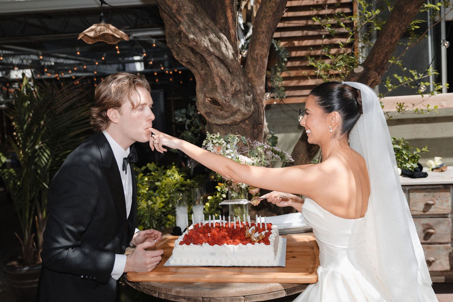 Bride in white gown feeds wedding cake to groom in black suit at outdoor reception with lush greenery.