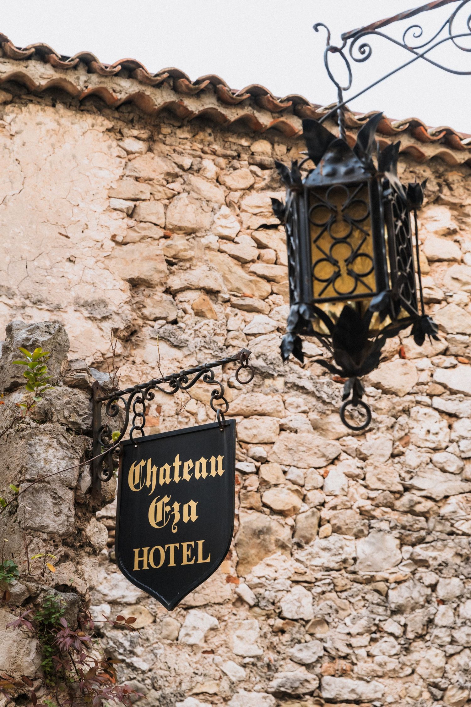 Château Eza - Black wrought iron sign for Chateau Eza Hotel mounted on rustic stone wall with ornate lantern in France.