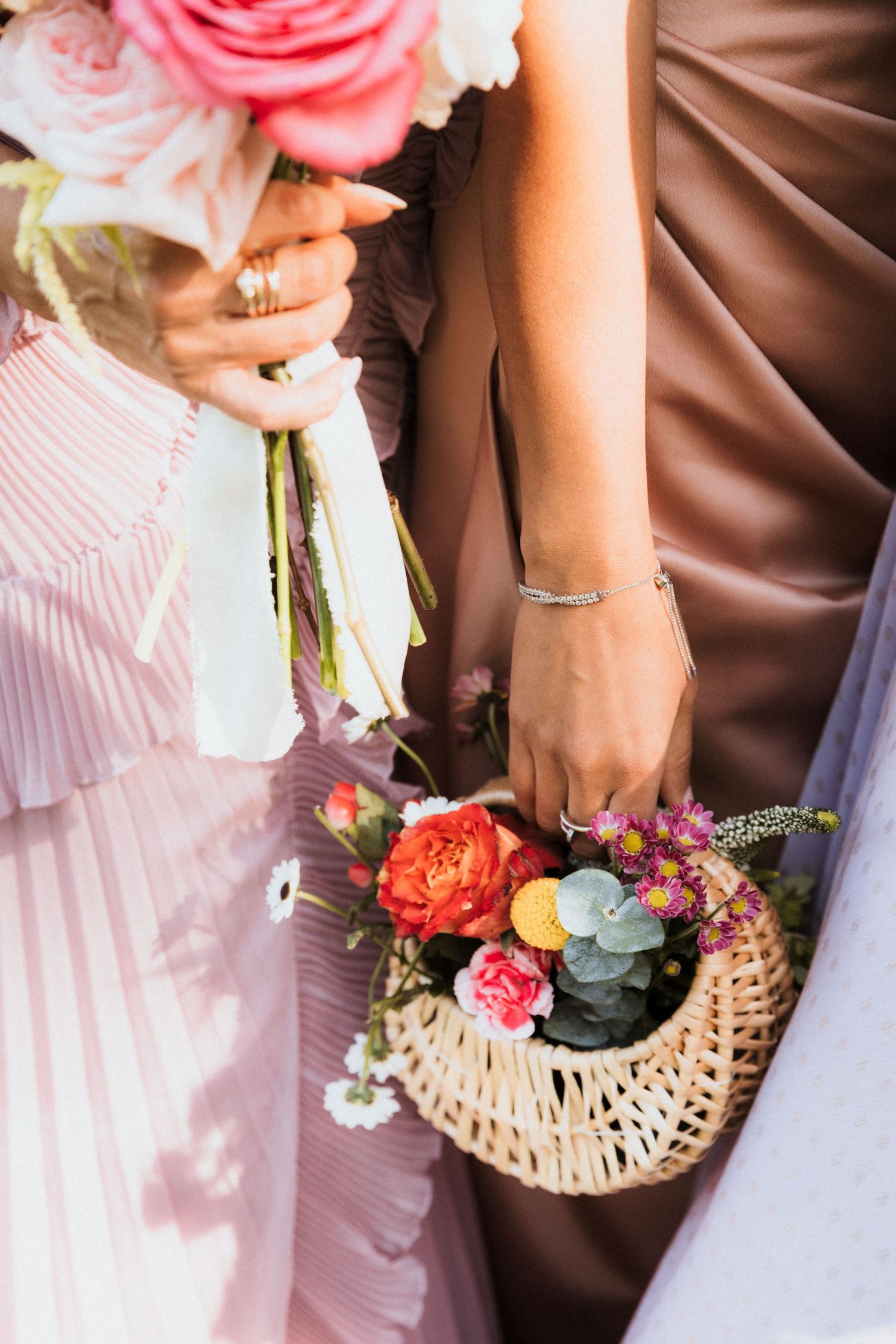 Two bridesmaids holding colorful floral bouquets, one in a wicker basket, wearing pink and brown dresses at a wedding.