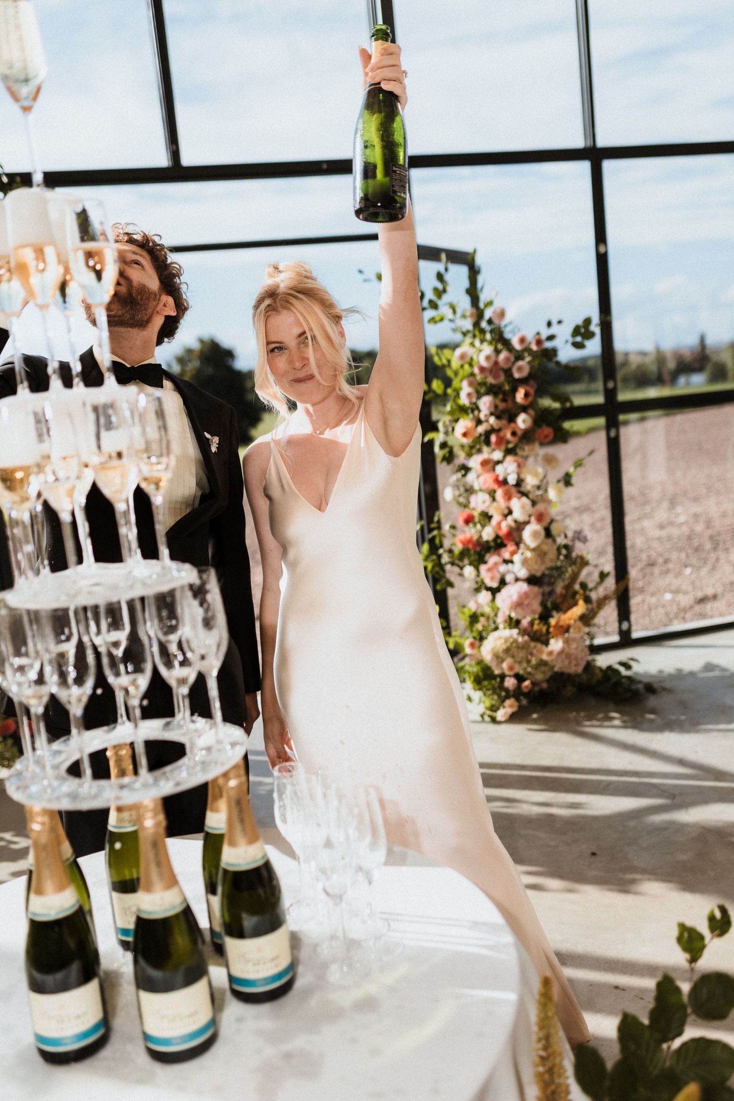 Bride in white slip dress raises champagne bottle beside a glass tower at a sunlit wedding reception.