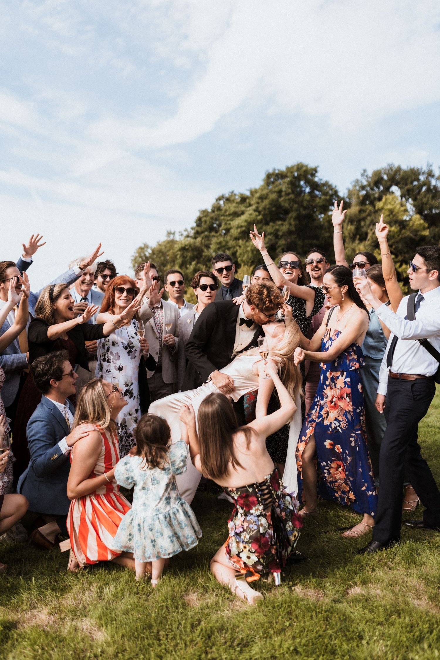 Excited wedding guests celebrate outdoors, cheering and posing together in a lively group photo on a sunny day.