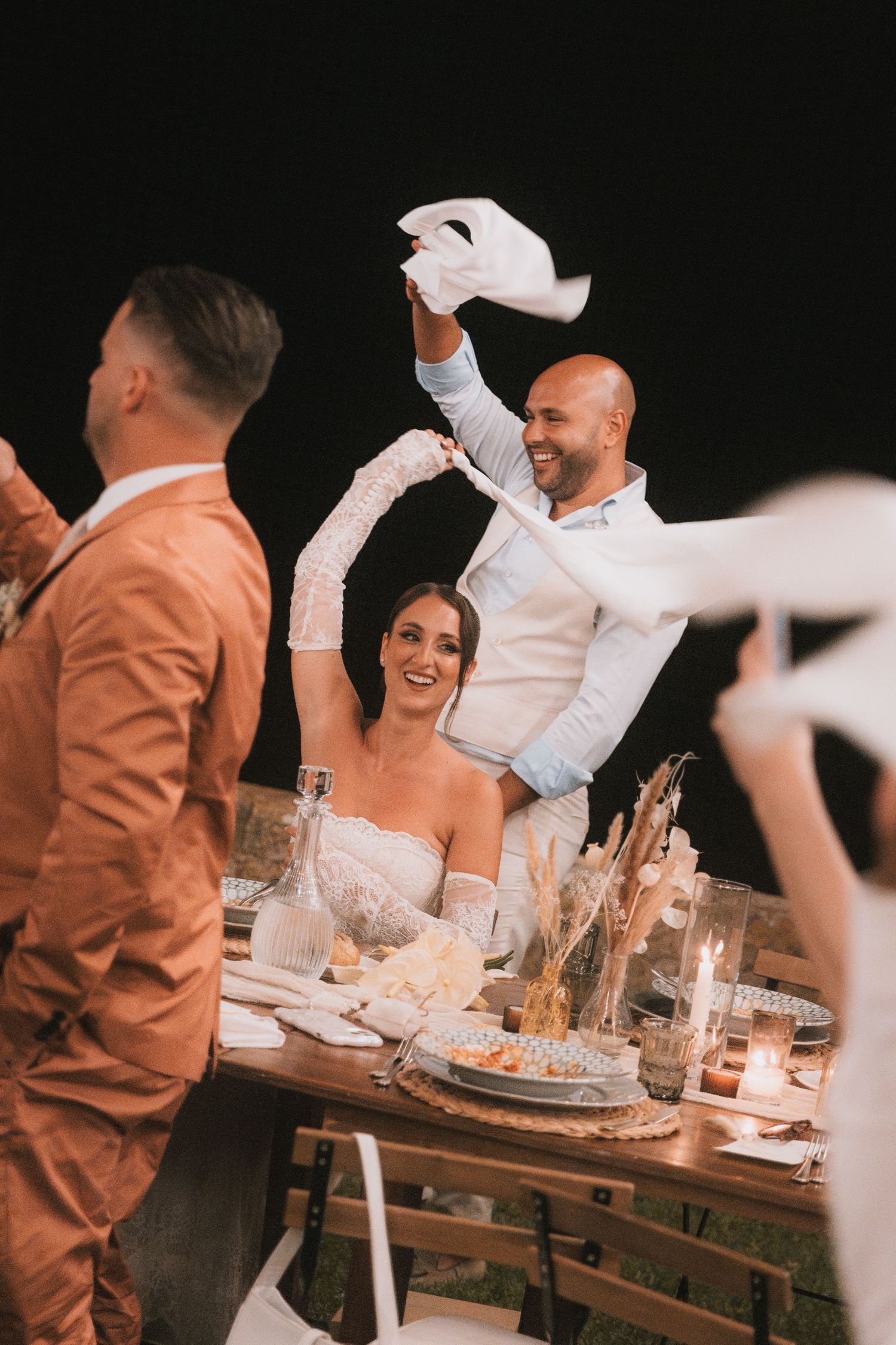 Bride and groom celebrate at outdoor wedding reception table, waving white napkins joyfully at night.