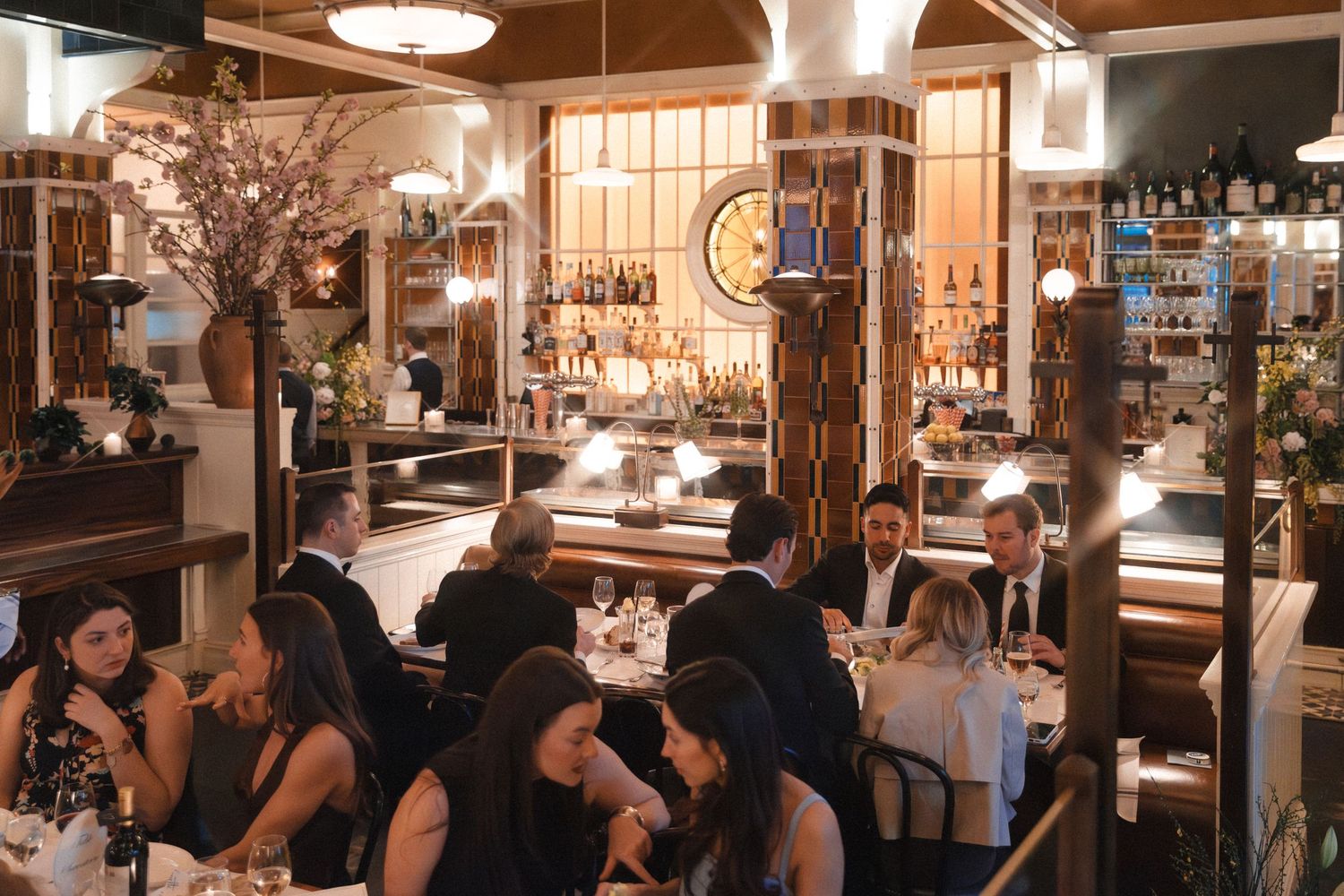 Elegant restaurant interior with guests dining at white-clothed tables, a glowing bar backdrop, and warm ambient lighting.