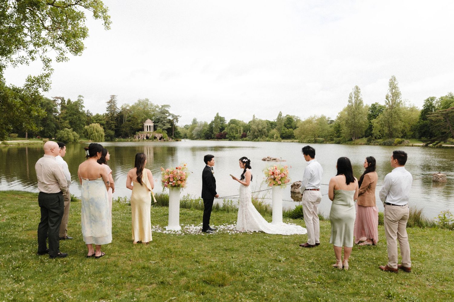 Outdoor wedding ceremony by a lake with couple at altar, guests in elegant attire standing on green lawn.