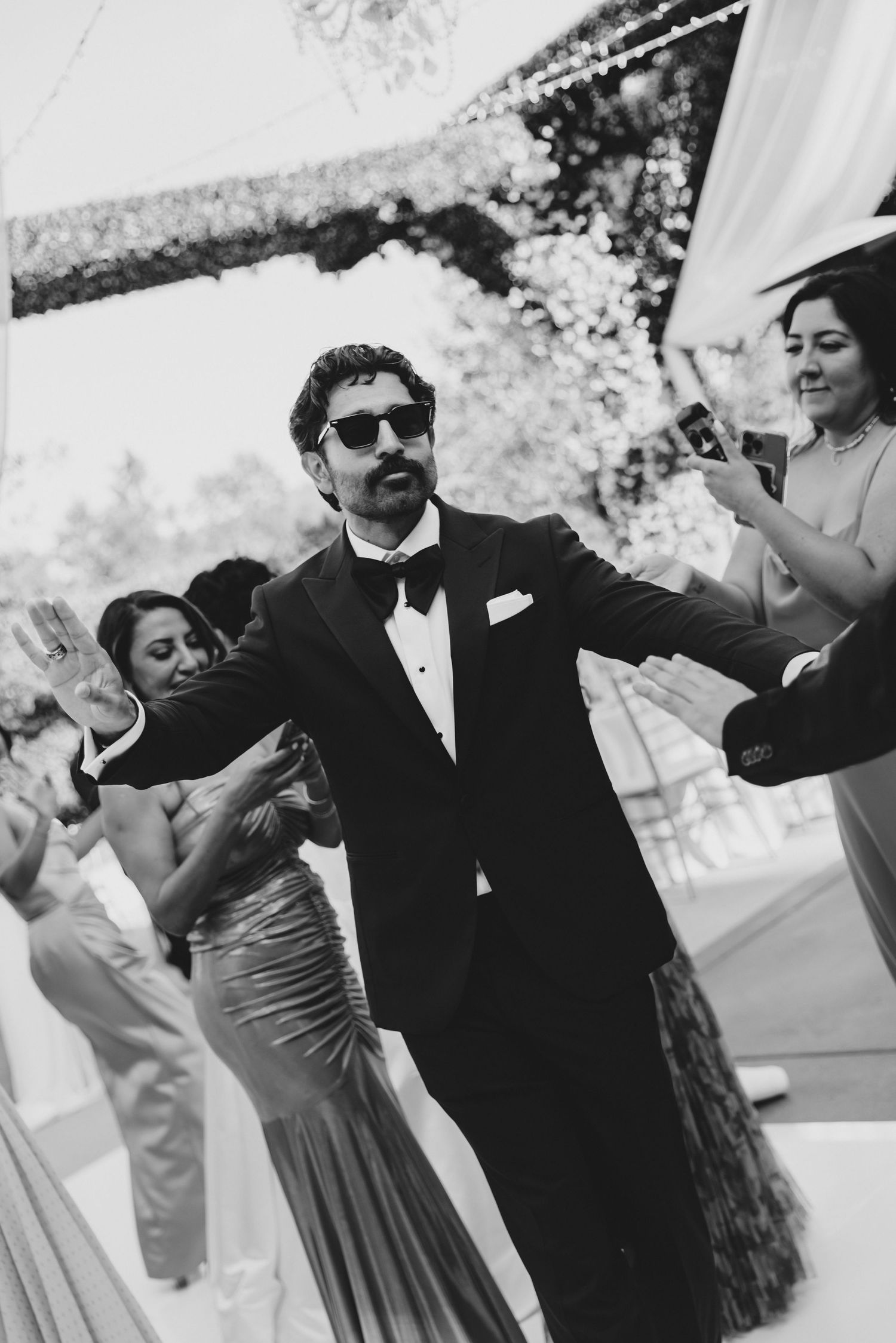 Groom in tuxedo and sunglasses dancing joyfully with guests at an elegant outdoor wedding reception.