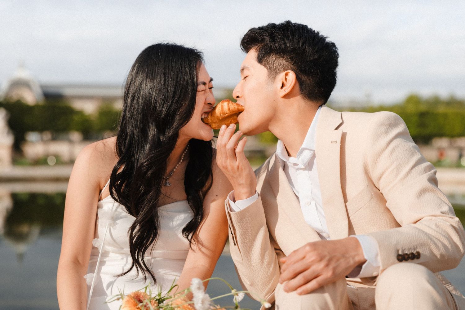 A couple in wedding attire shares a croissant kiss near a fountain in Paris.