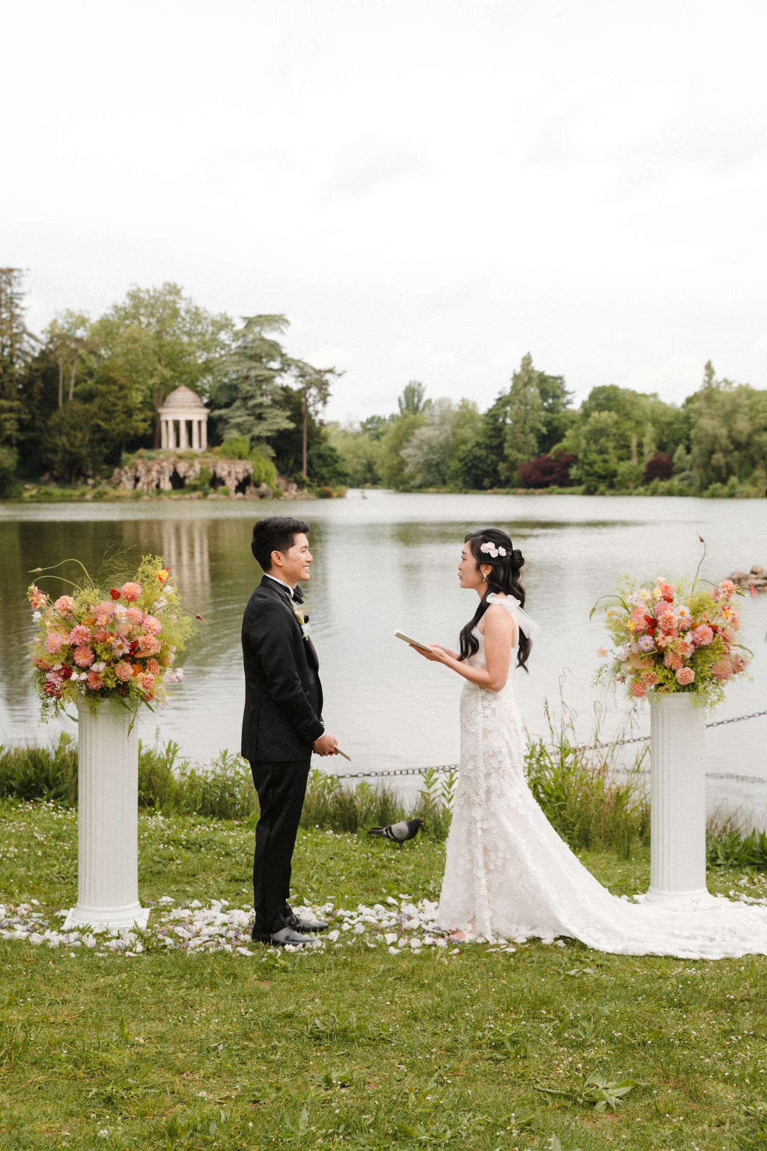 Couple exchanging vows outdoors by a lake, bride in lace gown, groom in tuxedo, flanked by floral arrangements on white pillars.