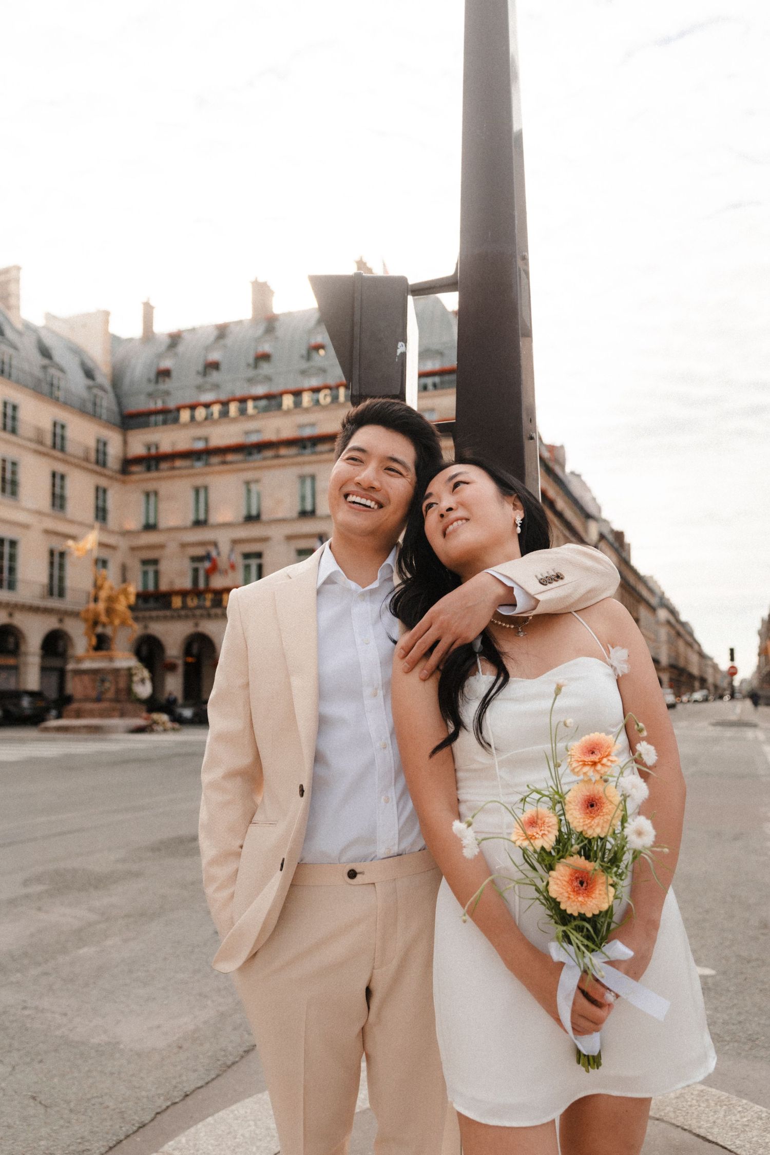 Happy Asian couple in wedding attire posing in Paris plaza, bride holding orange bouquet, groom in beige suit.