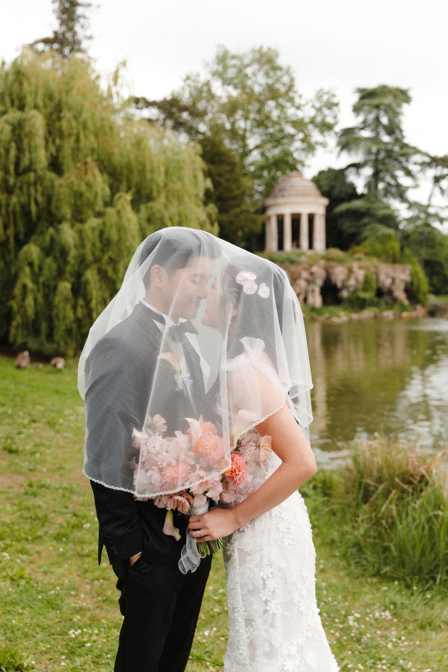 Bride and groom kissing under a veil by a pond with a classical temple gazebo in a lush garden.