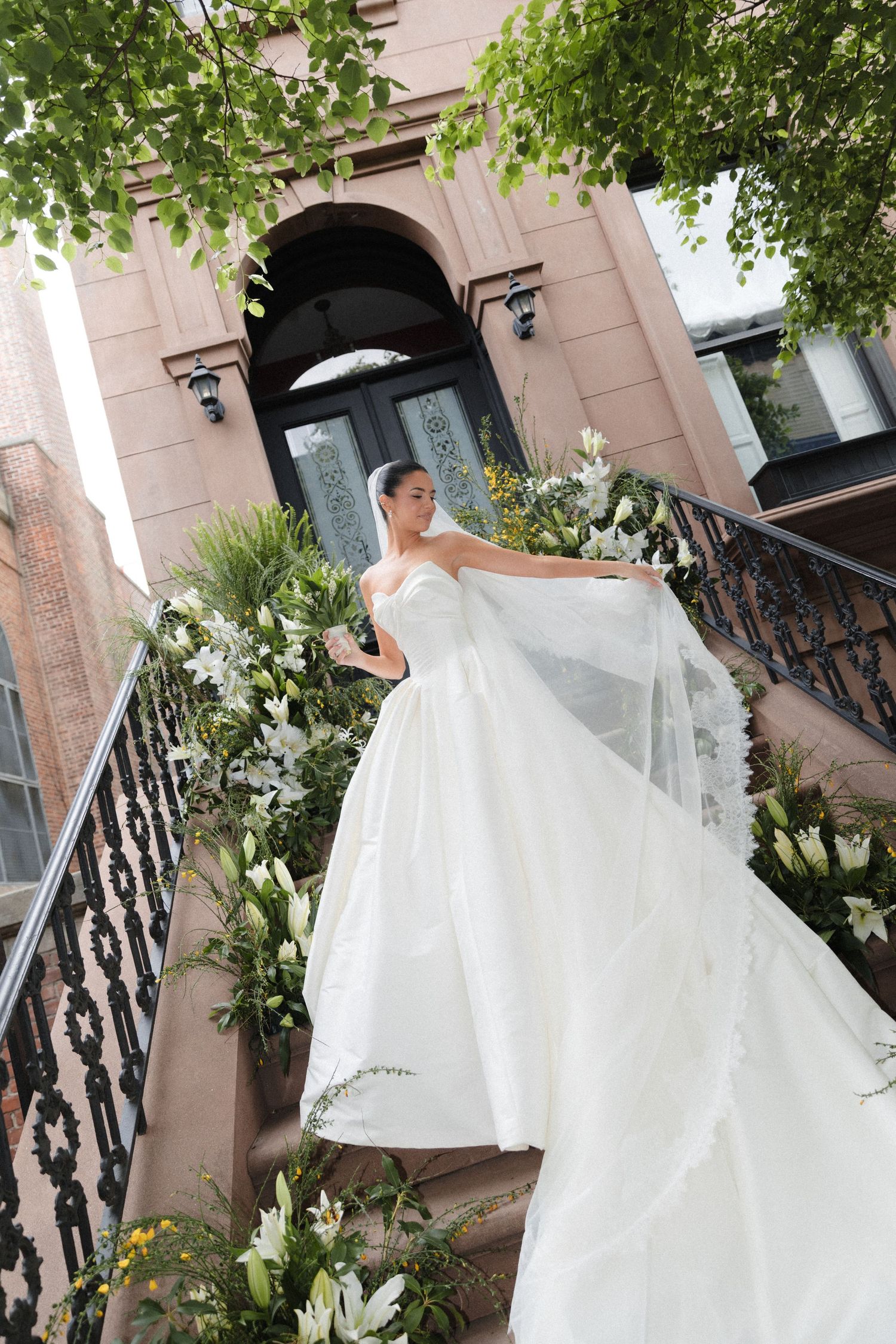 Bride in white strapless ball gown holding veil on floral-decorated staircase outside elegant pink building.