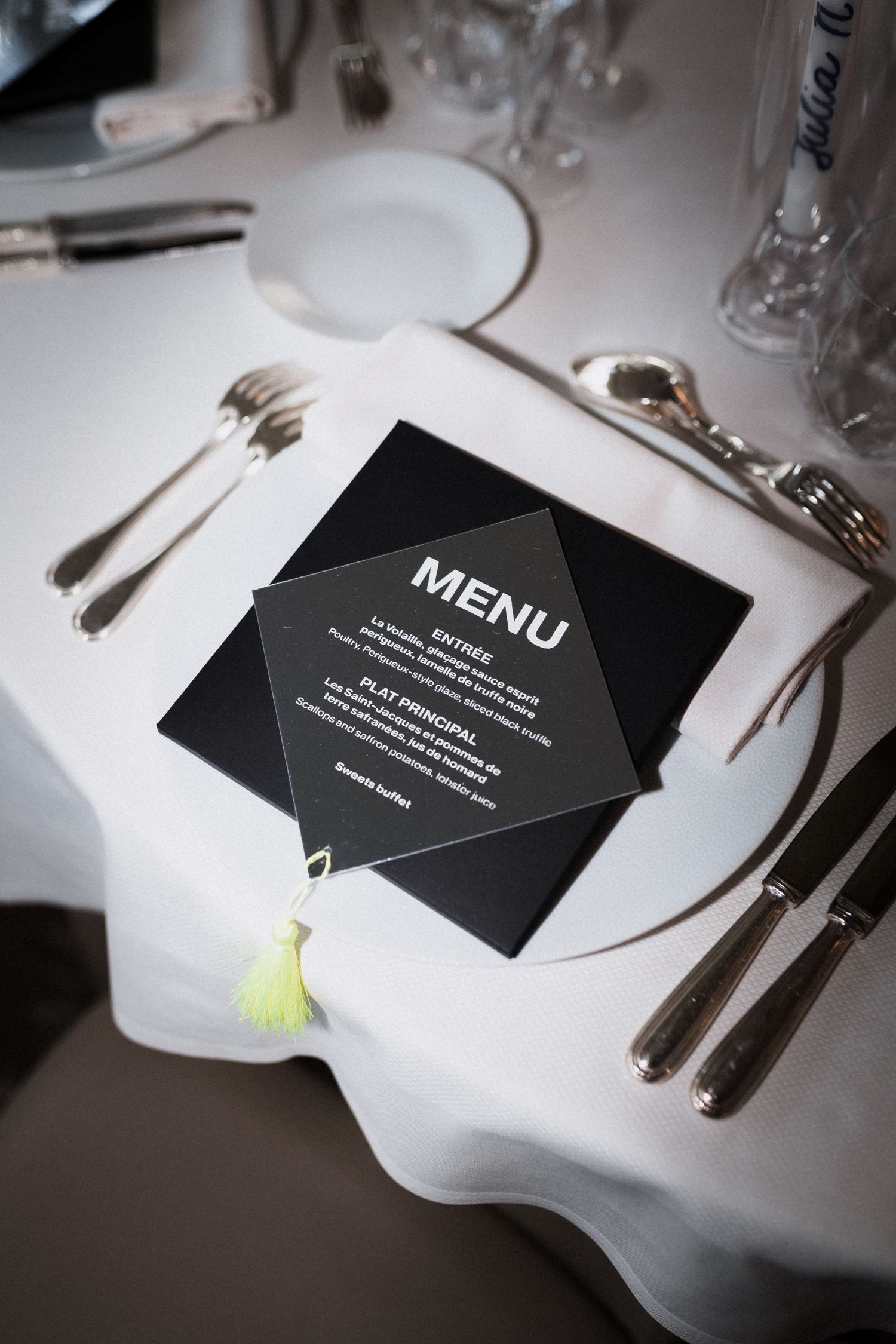 Black menu card placed on elegant white tablecloth with formal place setting and silverware at upscale restaurant.