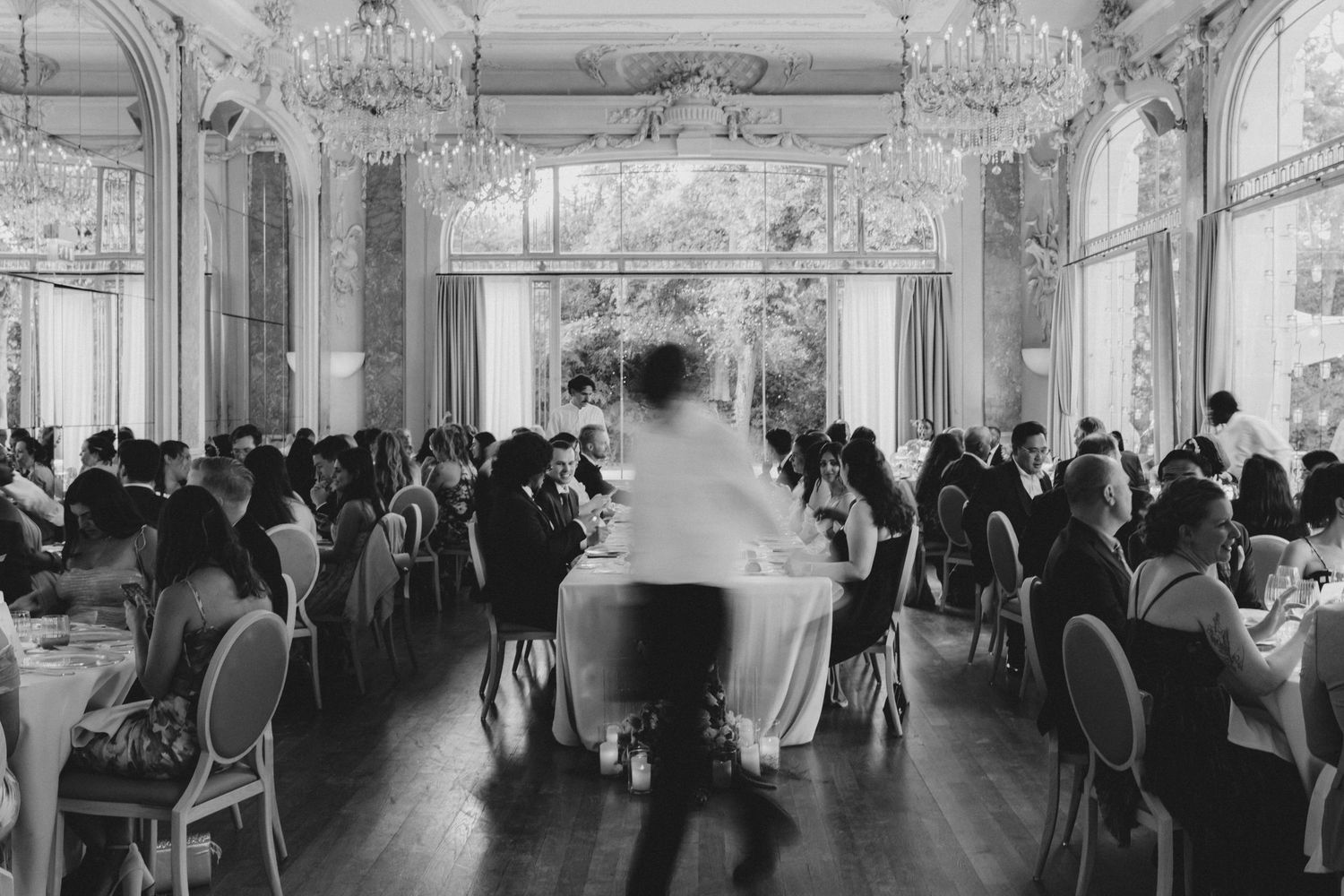 Black and white photo of elegant wedding reception in ornate ballroom with chandeliers, guests seated at tables.